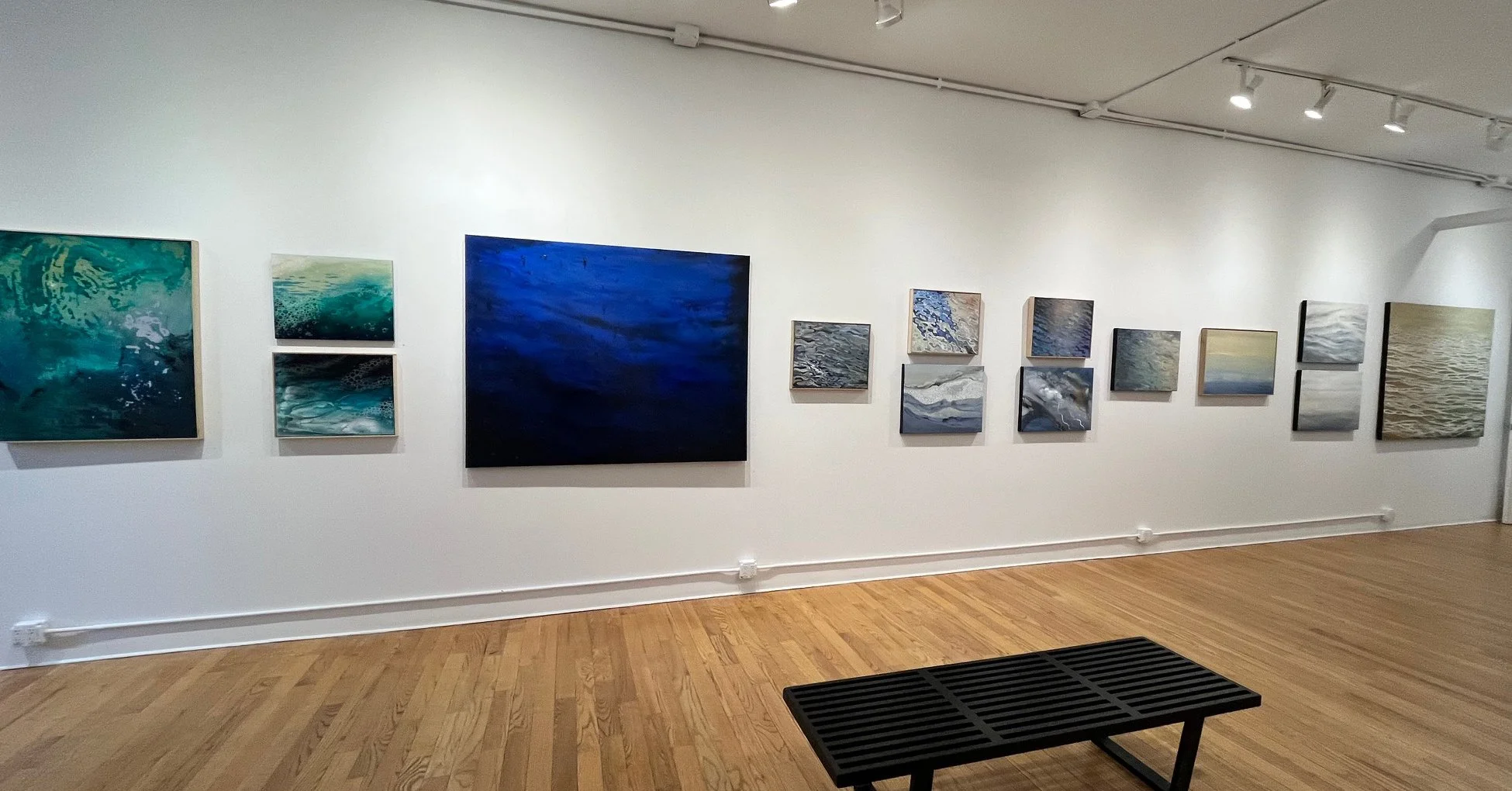 Gallery wall with several abstract paintings depicting water in various shades of blue, gray, and beige, arranged in a salon style in an art gallery with a hardwood floor and white walls.