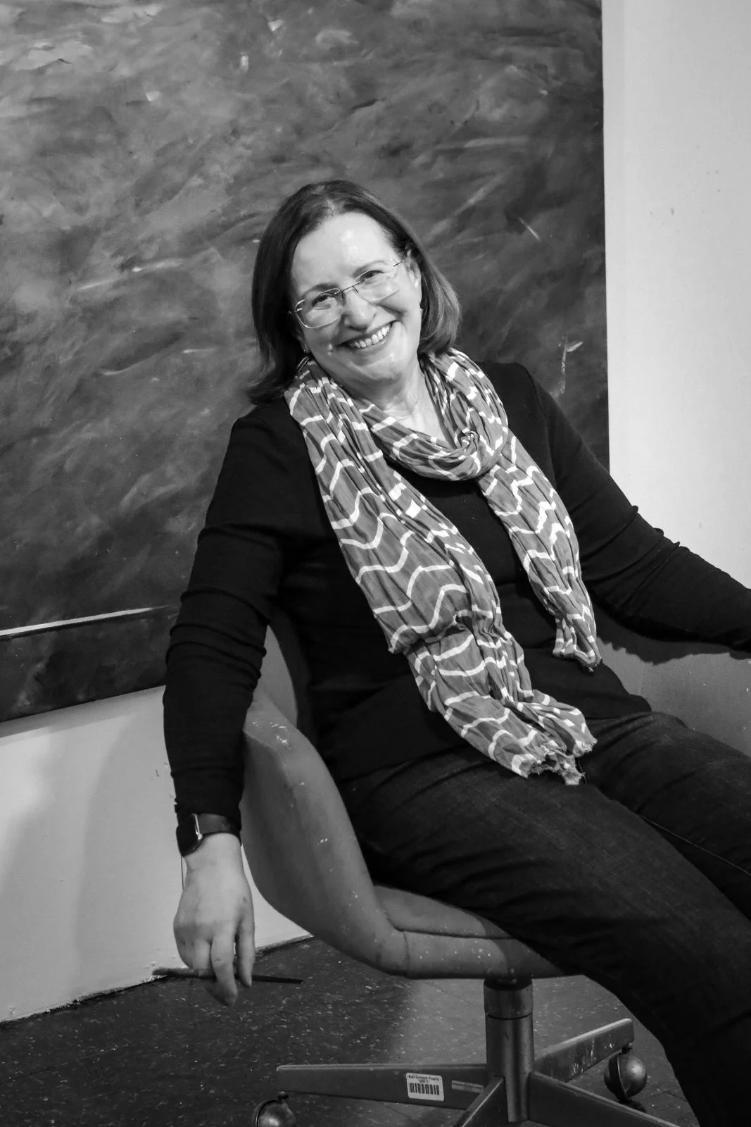 Smiling woman with glasses sitting on an office chair in front of an abstract painting in grayscale.