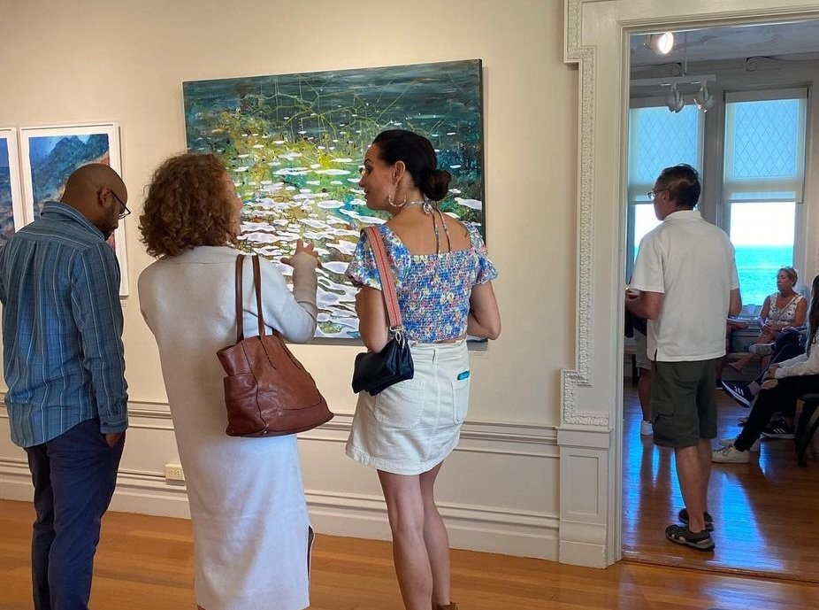 Group of people viewing and discussing artwork in an art gallery, with paintings on the walls and a window showing the outside.