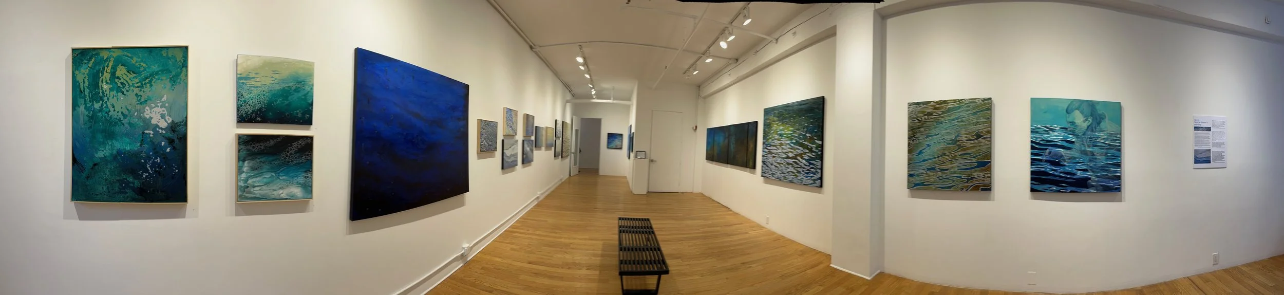 Inside an art gallery with abstract ocean-themed paintings on white walls. The paintings are mostly blue, green, and gold. Wooden floor and a black bench are visible in the center.