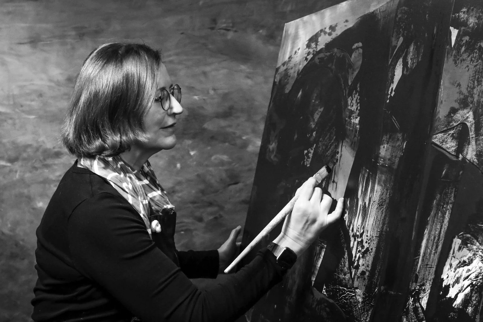 A woman with shoulder-length hair and glasses painting on a large canvas in an art studio.