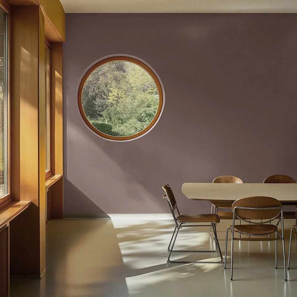 Flügger AI generated scene by Scenes. Minimalist dining area with round window, brown wall, modern table and chairs, and view of greenery outside.