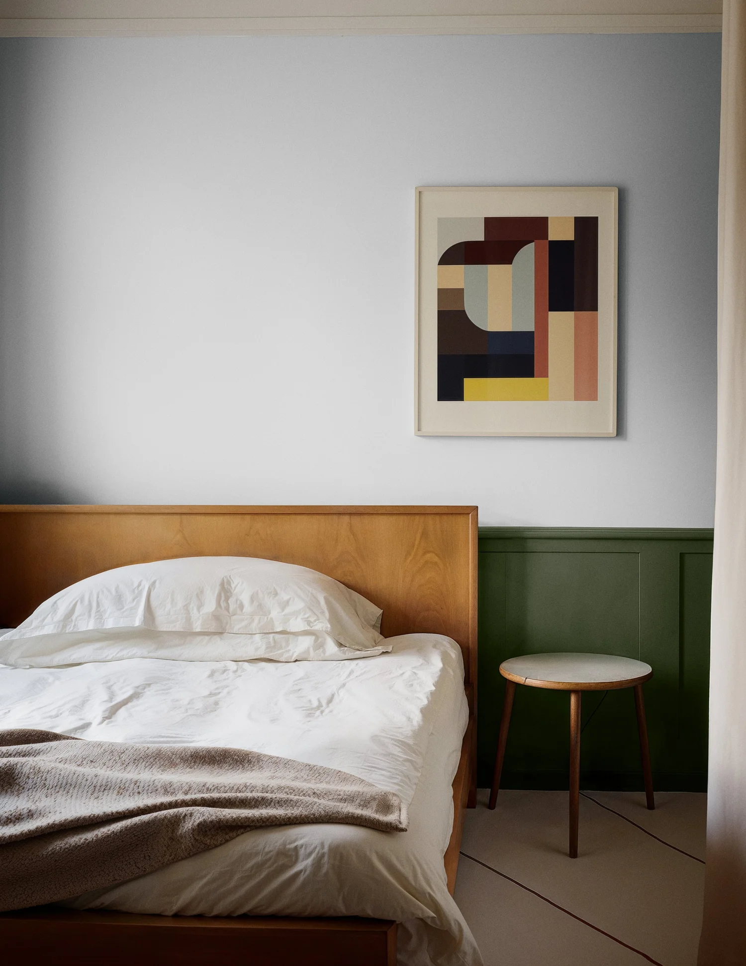 Minimalist bedroom with wooden bed frame, white bedding, green panel wall, abstract artwork, and round wooden side table.