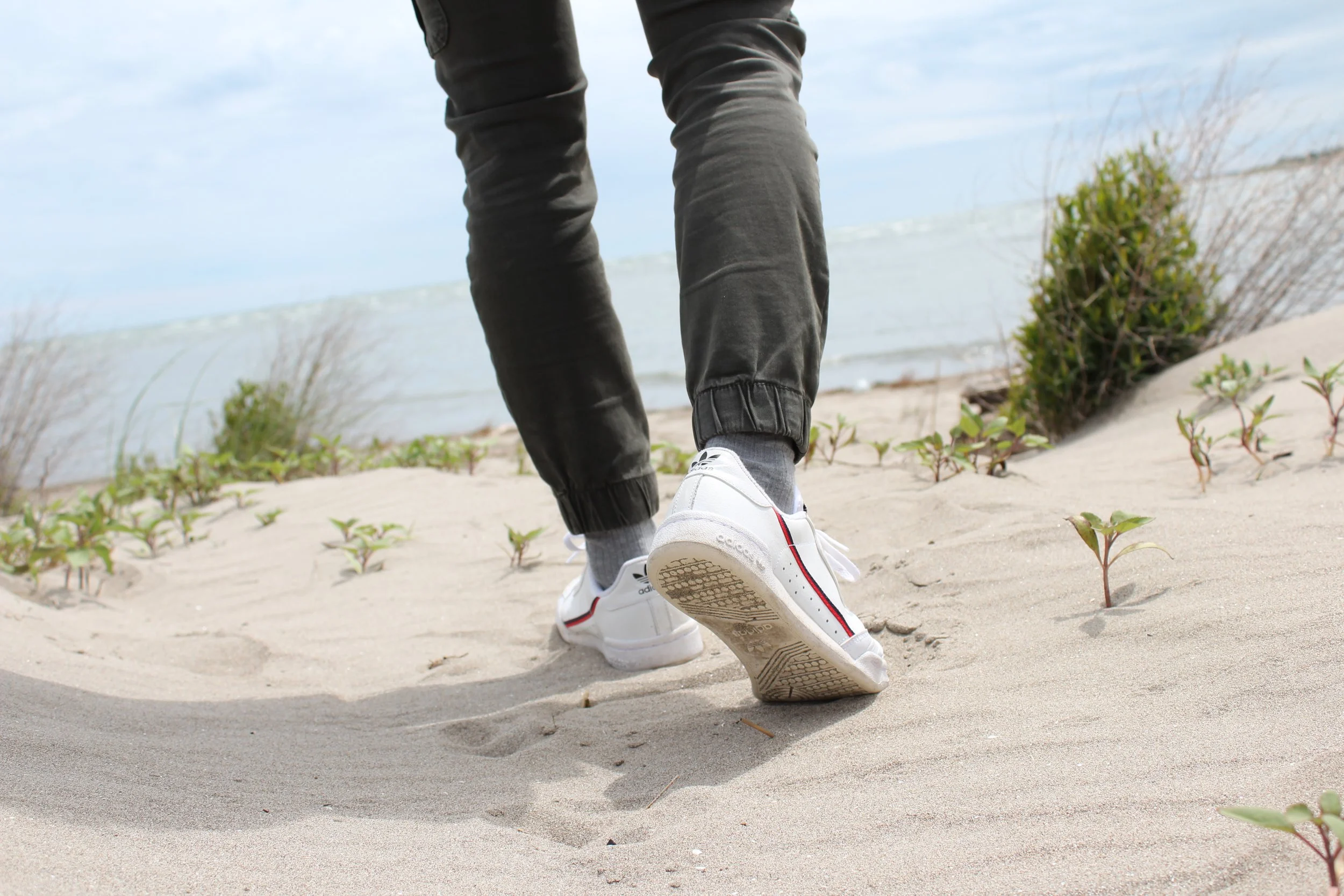 Persona che cammina sulla sabbia vicino al mare, con scarpe sportive bianche e pantaloni scuri. Sullo sfondo, vegetazione e cielo nuvoloso.