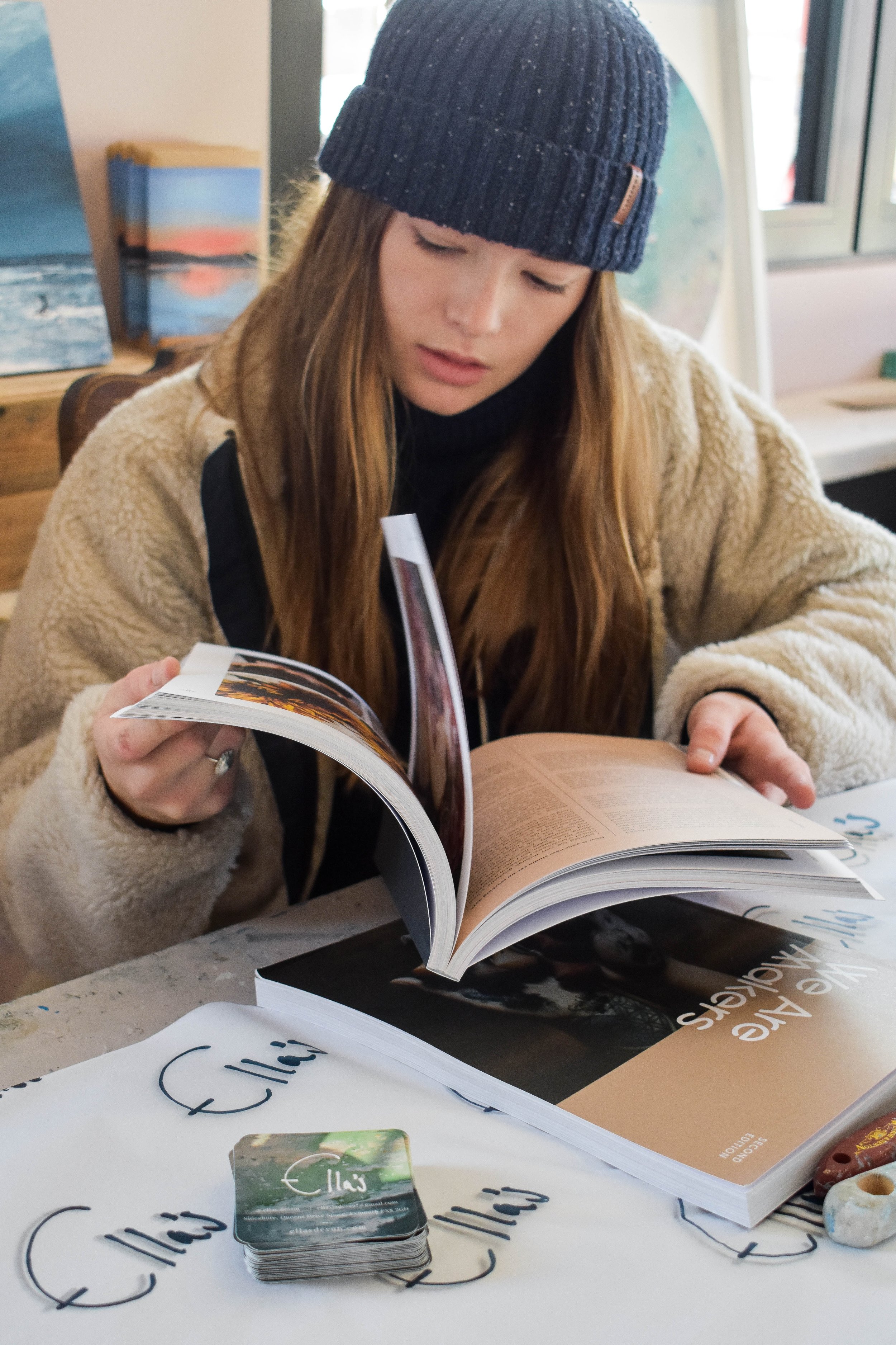 Persona con cappello blu legge un libro aperto su un tavolo con biglietti da visita e un'altra rivista.