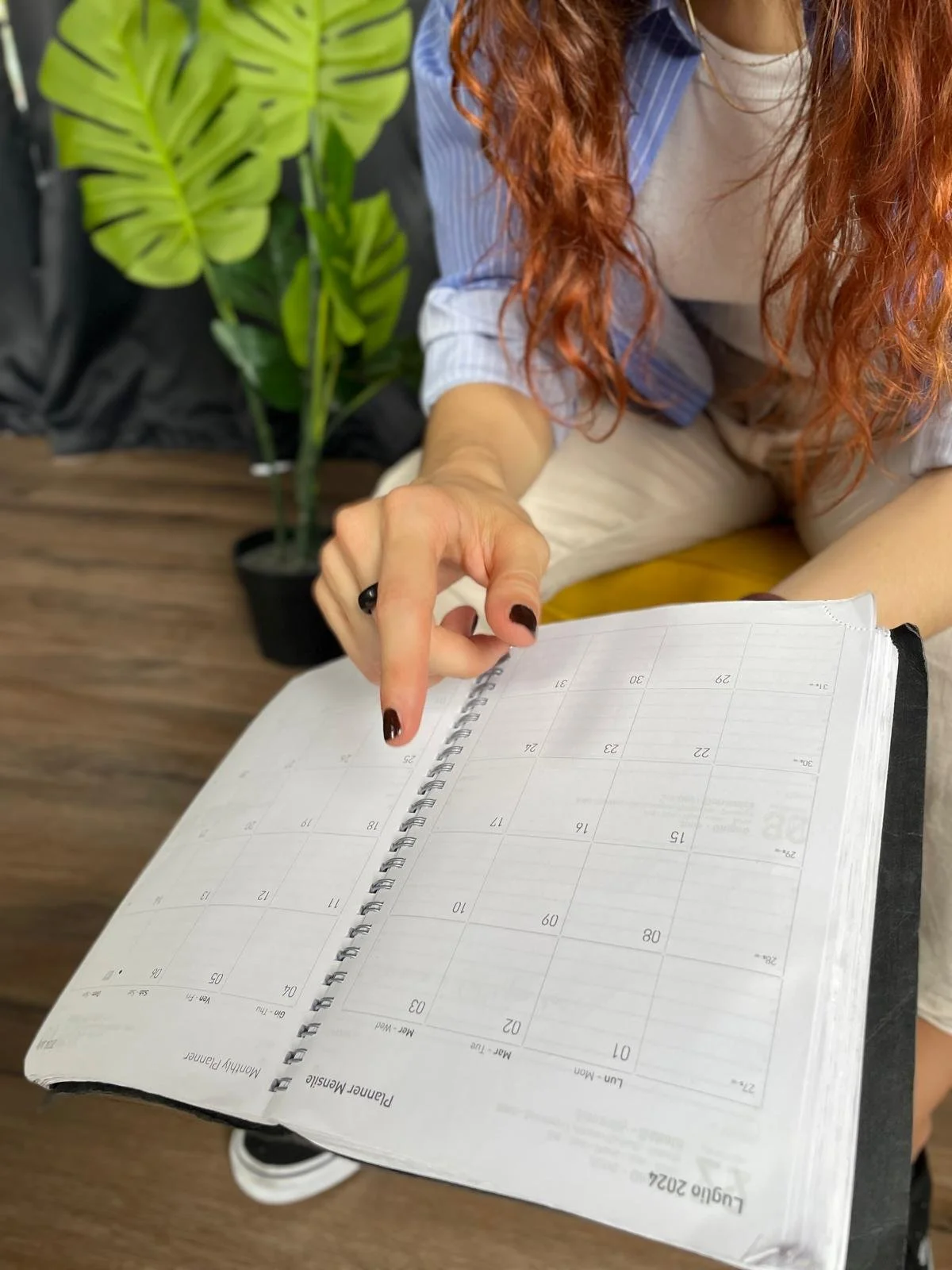 Donna con capelli rossi che indica un calendario aperto, seduta accanto a una pianta d'appartamento, sfondo di un ambiente interno.