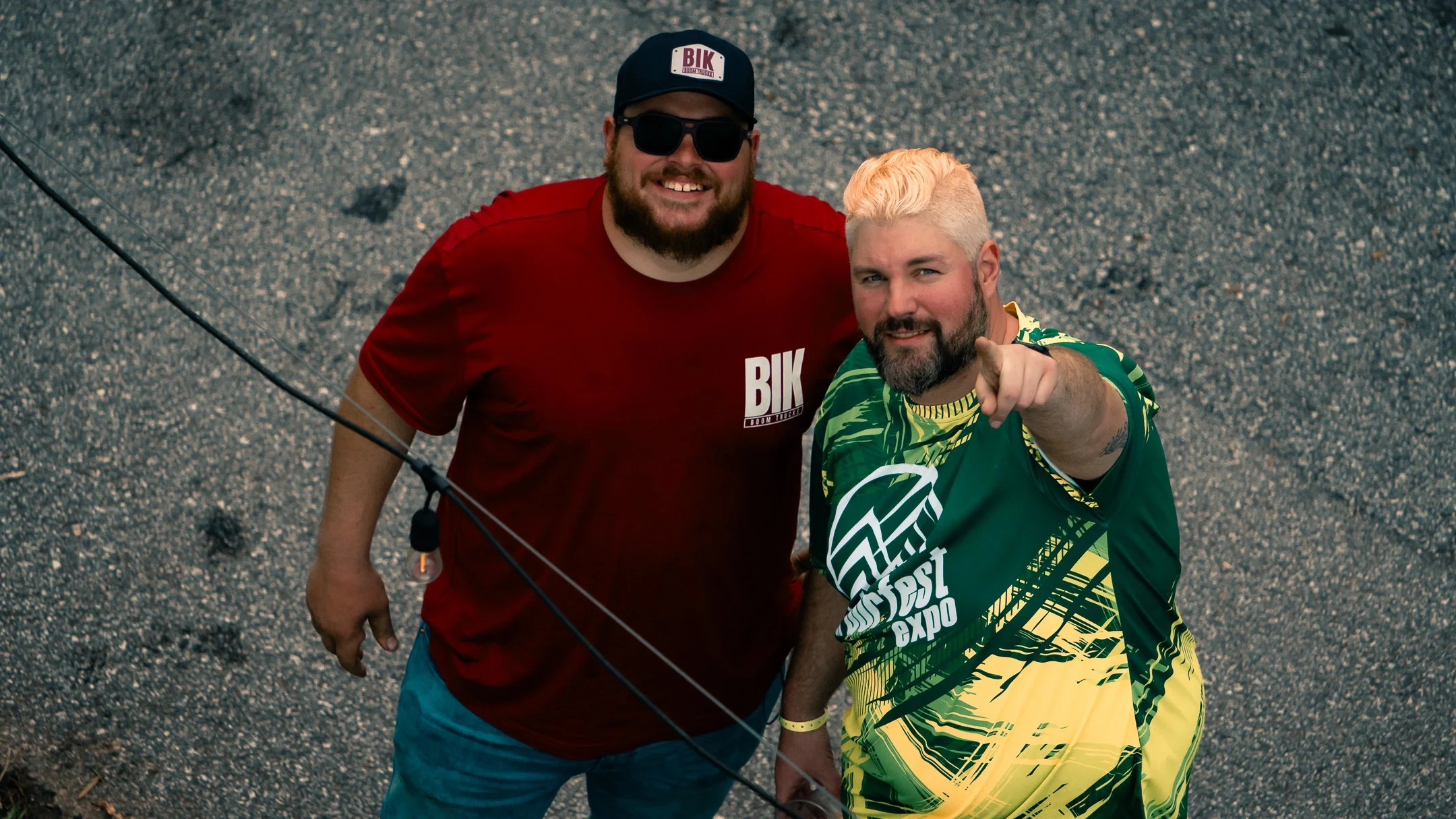 Two smiling men looking up at the camera. One has a beard, sunglasses, a black cap, and a red shirt with "BIK" written on it. The other has a beard, platinum blonde hair, and a green and yellow shirt with graffiti patterns. The man in the graffiti shirt is pointing at the camera. They are standing on a gravel surface.