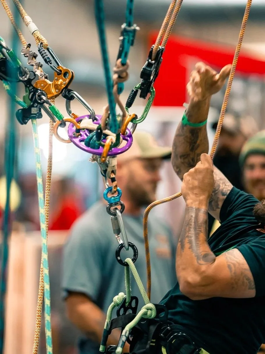 Climbers' quickdraws, carabiners, and climbing ropes hanging on a gear rack with people in the background at an indoor climbing gym.
