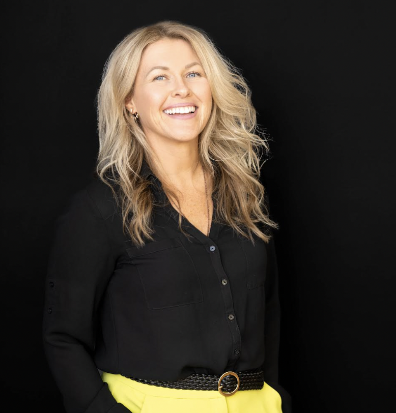 Smiling woman with long blond hair, wearing a black shirt and yellow pants, standing against a black background.