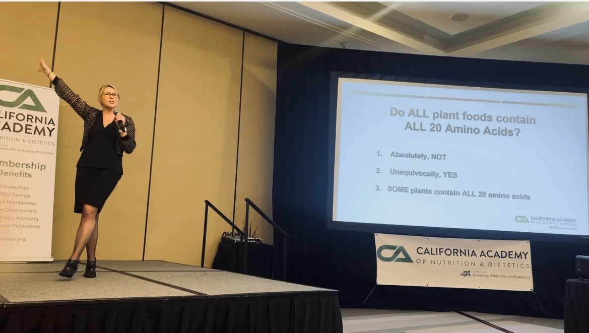 Speaker presenting on stage with a slide displaying the question 'Do ALL plant foods contain ALL 20 Amino Acids?' Options are: 1. Absolutely, NOT 2. Unequivocally, YES 3. SOME plants contain ALL 20 amino acids. California Academy of Nutrition & Diete