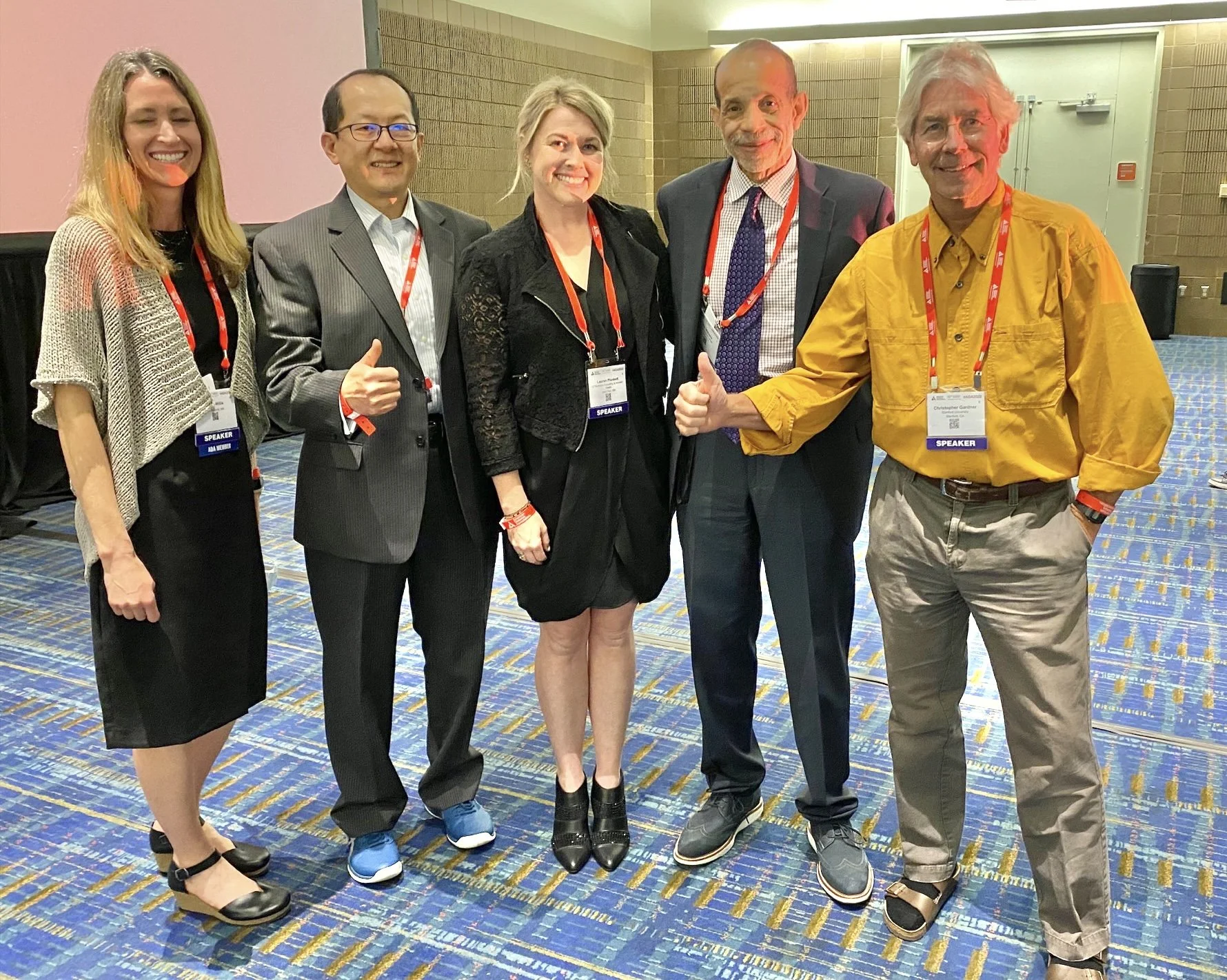 A group of five people standing in a conference room, all wearing lanyards with "SPEAKER" badges. They appear to be posing for a photo, with some giving thumbs up. The room has a blue patterned carpet and a presentation area in the background.