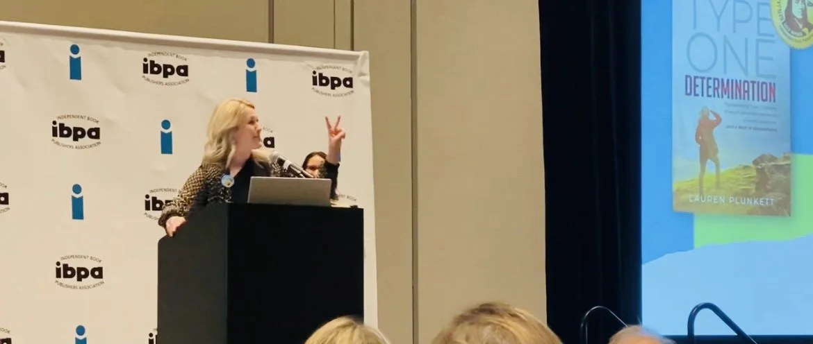 A woman speaking at a podium with a microphone at a conference. She is gesturing with her hand, making a peace sign. Behind her is an IBPA (Independent Book Publishers Association) banner. To the right, there's a large projection showing a book cover