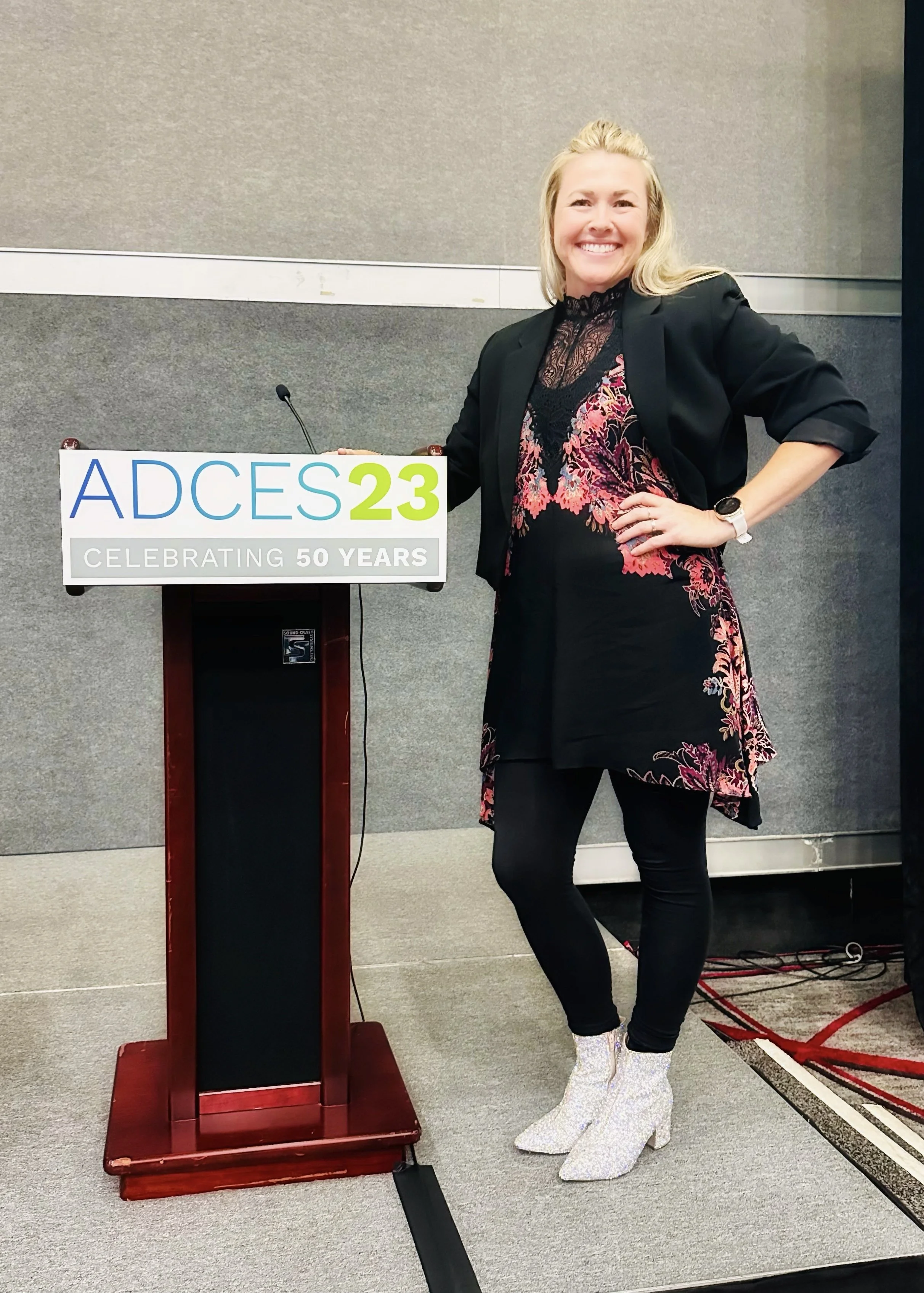 Person standing next to a lectern with ADCES23 sign, celebrating 50 years, wearing a black and floral outfit.