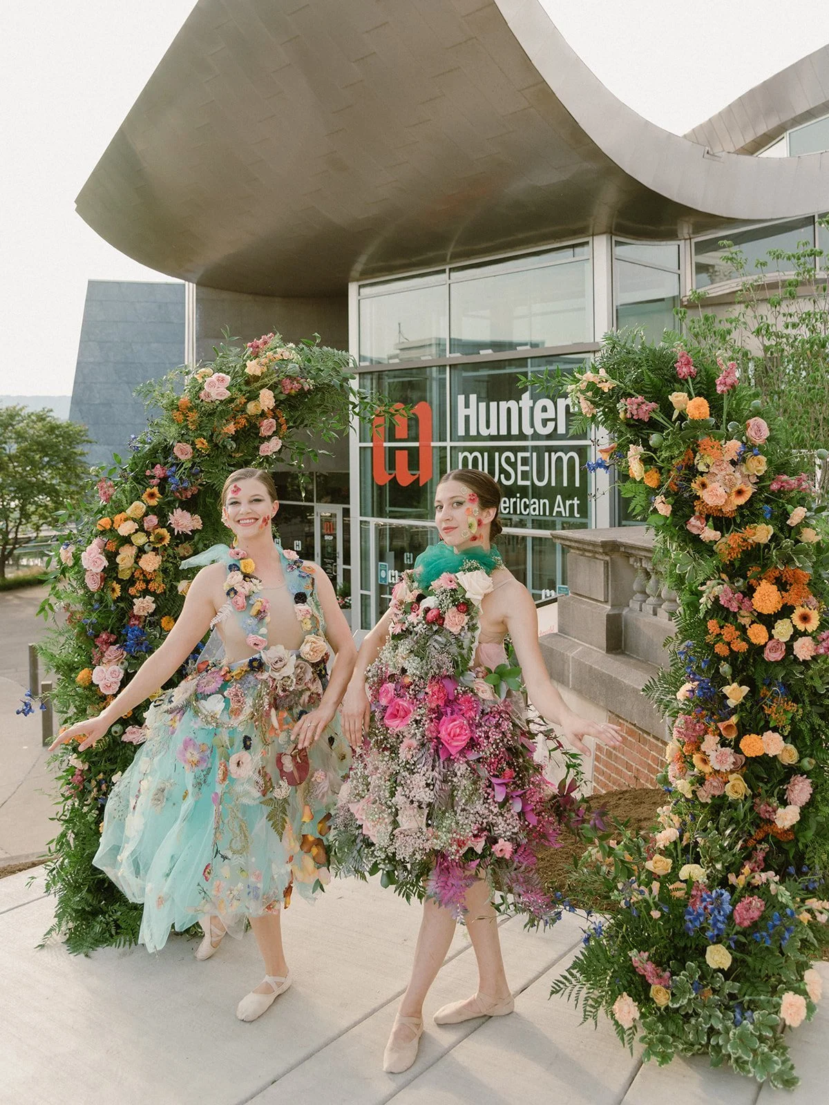 Two women dressed in floral costumes standing under a flower arch in front of the Hunter Museum of American Art.