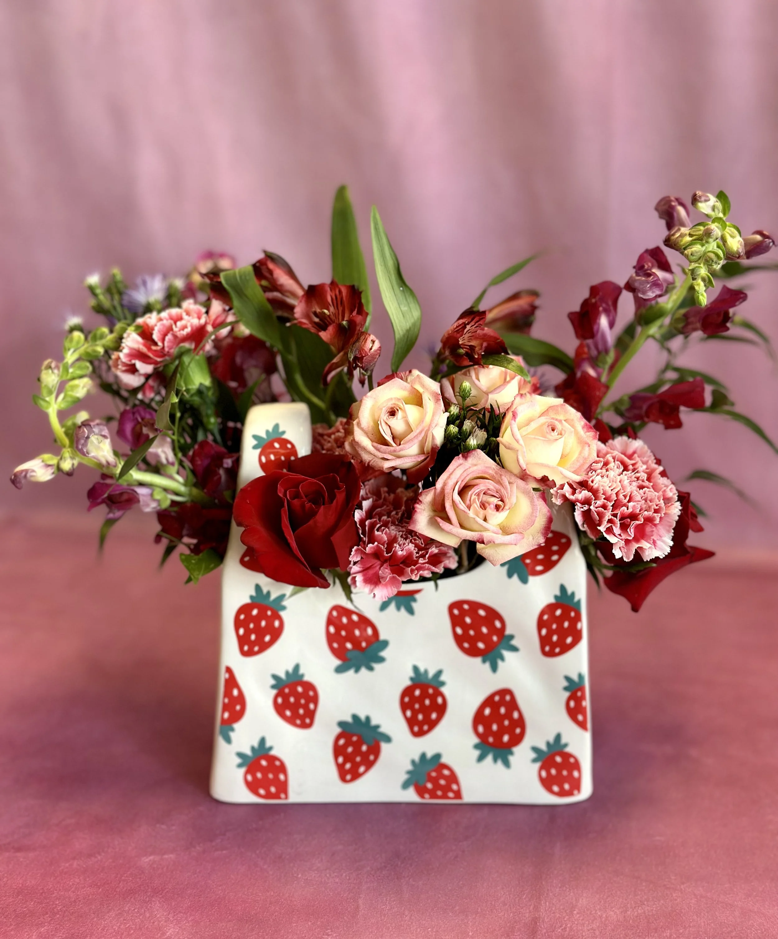 A floral arrangement in a white vase decorated with red strawberries, set against a pink background.