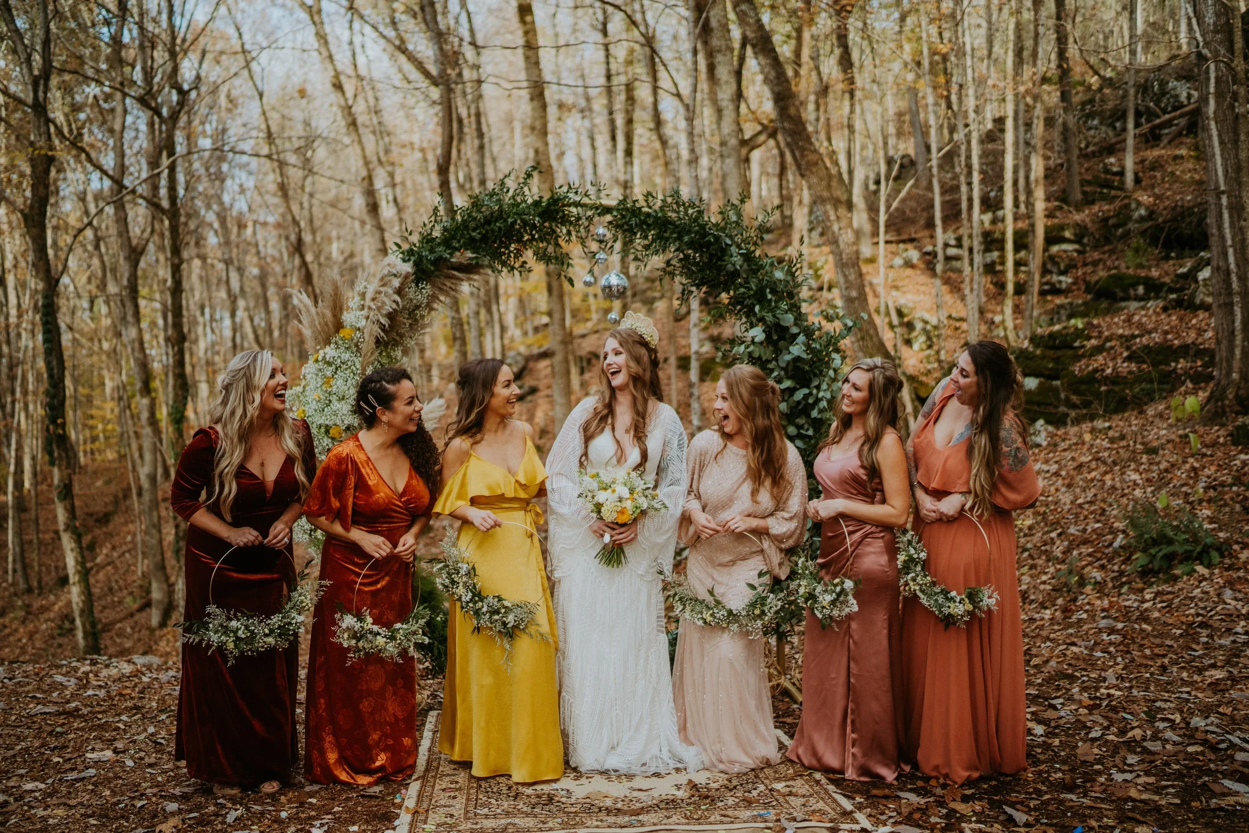 A bride in a white wedding dress with a bouquet stands among seven bridesmaids in colorful dresses, smiling and laughing under a decorated arch in a forest during fall.