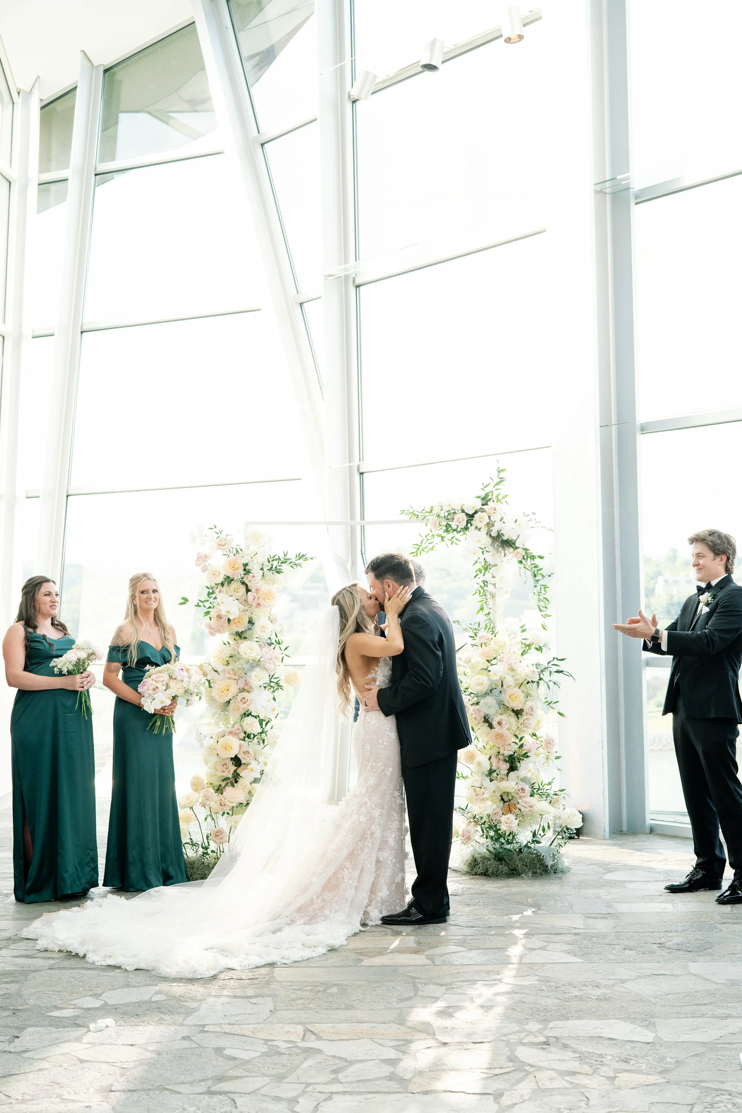 A bride and groom share a kiss during their wedding ceremony in a modern, glass-walled venue. The bride wears a white lace wedding gown with a long train, and the groom is in a black tuxedo. Two bridesmaids in green dresses and a groomsman clapping stand nearby. Floral arrangements with pastel pink and white flowers decorate the background.