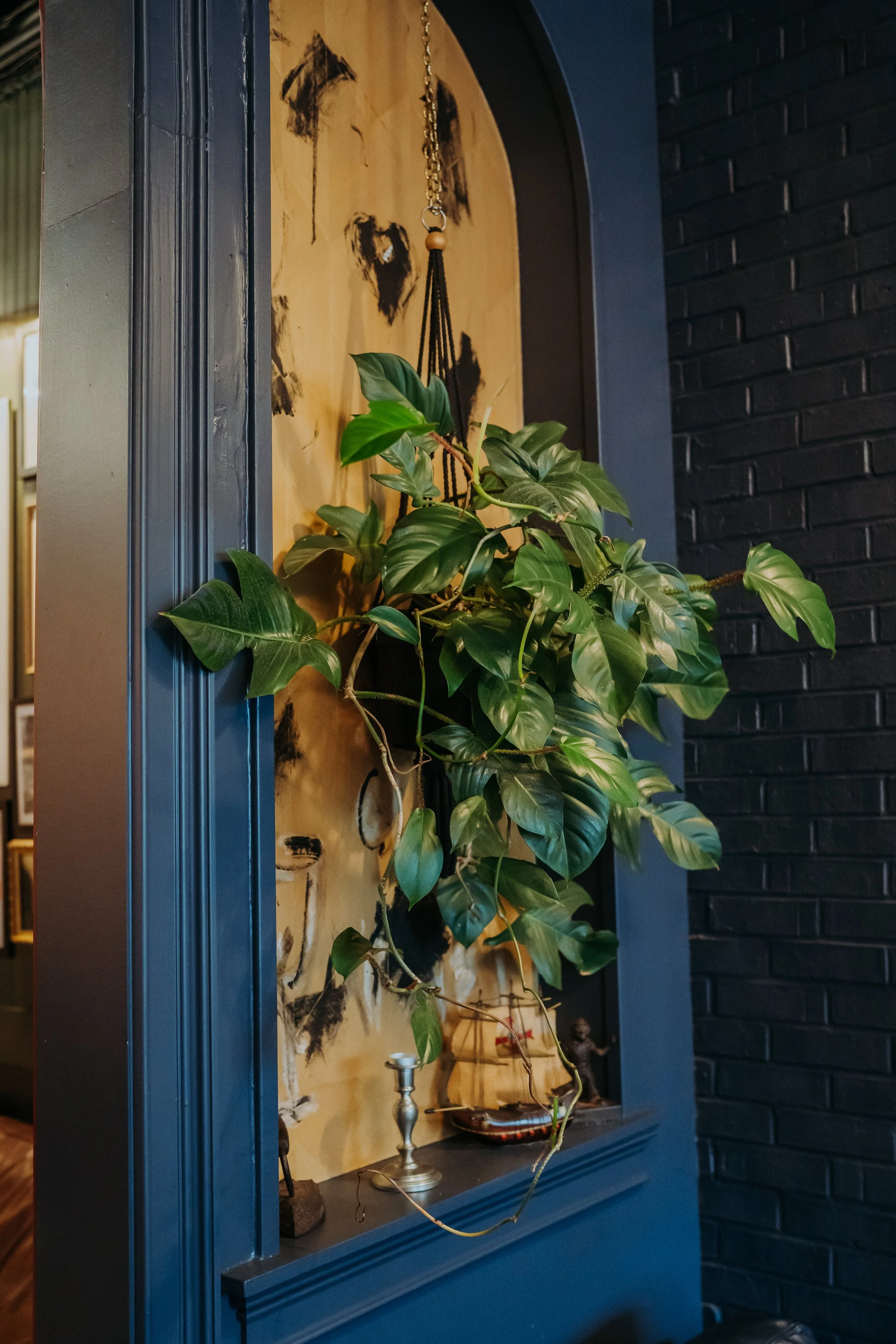 Decorative interior shelf with green hanging plant, small ship model, metal candlestick, and figurine, against a yellow wooden backing and surrounded by dark blue and black walls.