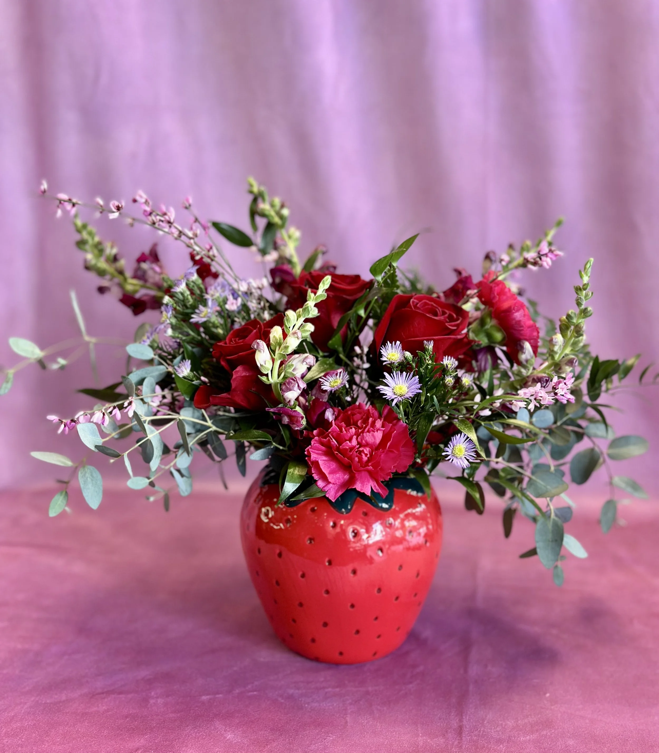 A flower arrangement with red roses, pink carnations, small purple daisies, and mixed greenery in a red strawberry-shaped vase on a pink background.