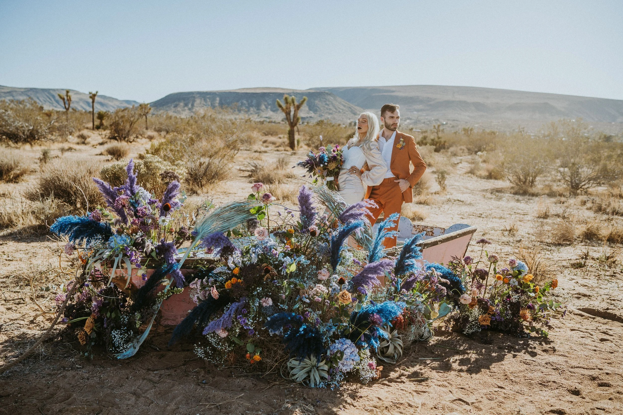 A bride and groom stand in a desert landscape, surrounded by colorful flowers, next to an old pink horse-drawn cart.