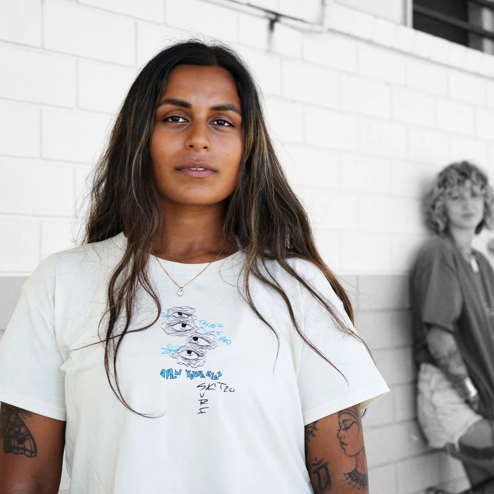 A woman with long dark hair and tattoos on her arms stands in front of a wall, with two women in the background leaning against the wall, one wearing a hat and the other with curly hair, on a sunny day.