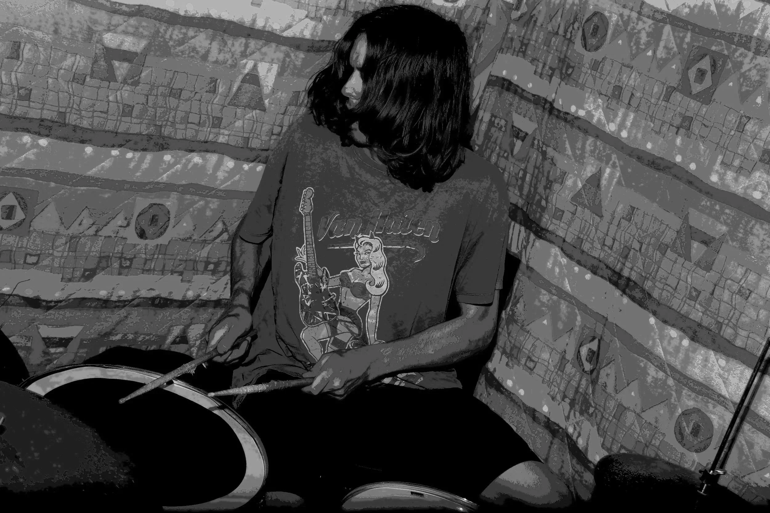 A person with shoulder-length dark hair wearing a Van Halen graphic t-shirt playing drums against a colorful, patterned backdrop.