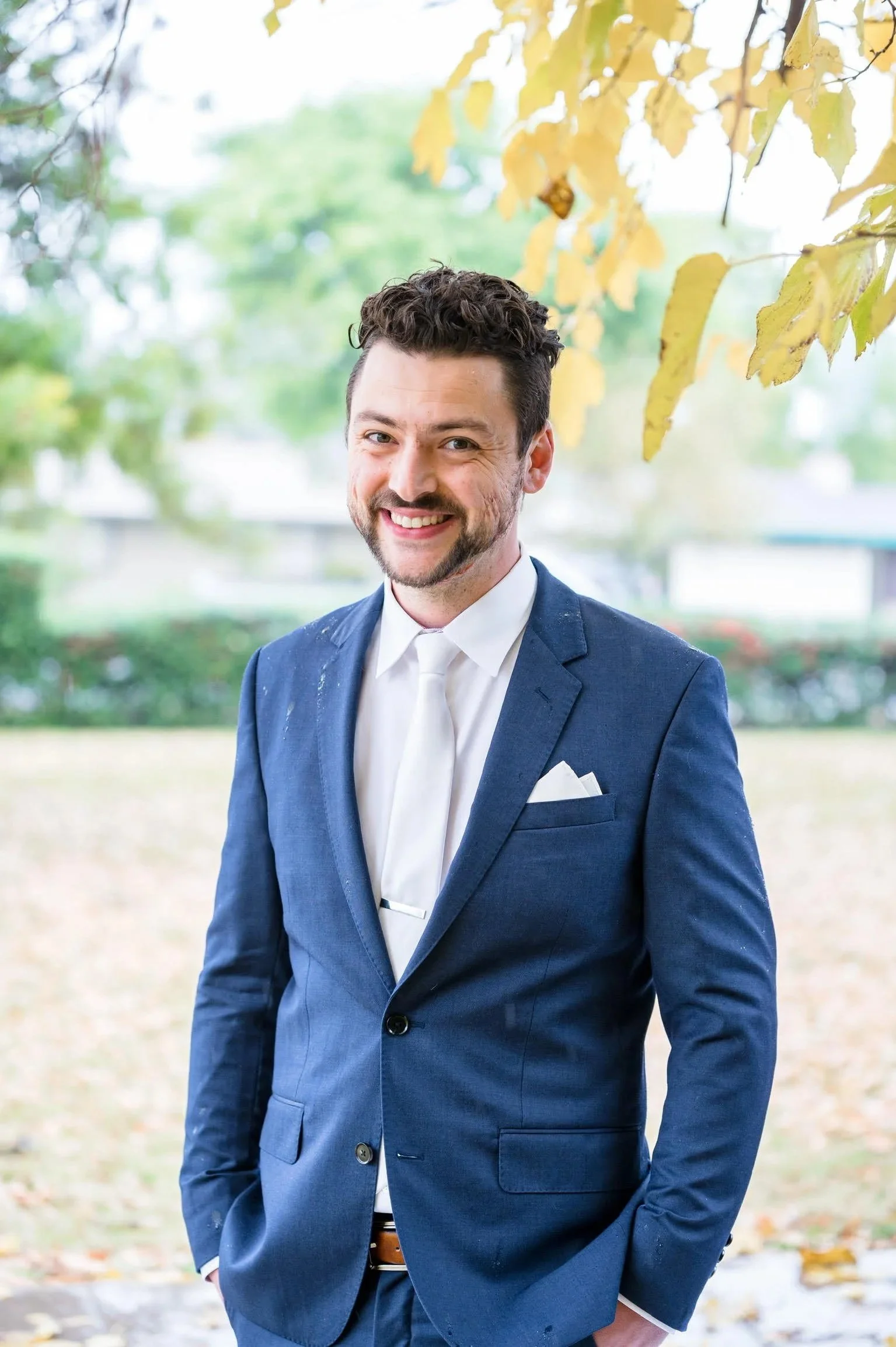 A portrait of a smiling man wearing a blue suit and white shirt standing outdoors with trees and yellow leaves in the background. Tobin Kopp, P.E. Civil Engineer