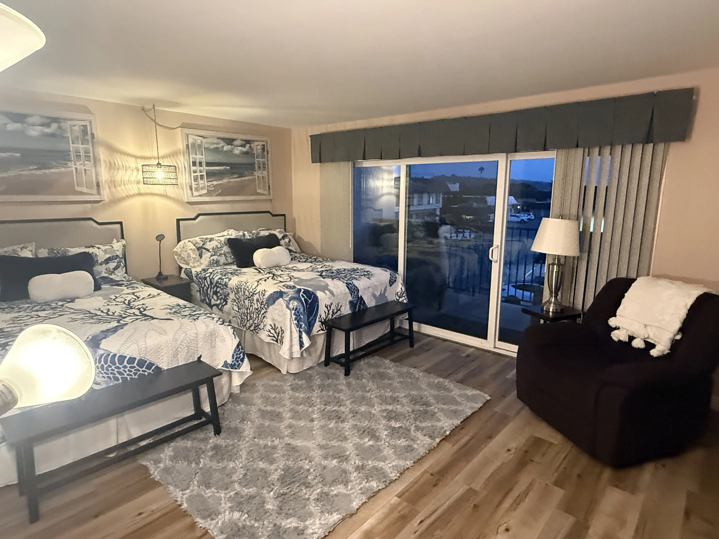 The Queens Suite bedroom in our Solana Beach vacation rental. A bedroom with two comfy queen-sized beds, an en suite, a walk-in closet with games and beach supplies, a plush rug, lamps, a La-Z-Boy, and a sliding glass door leading to a private balcon
