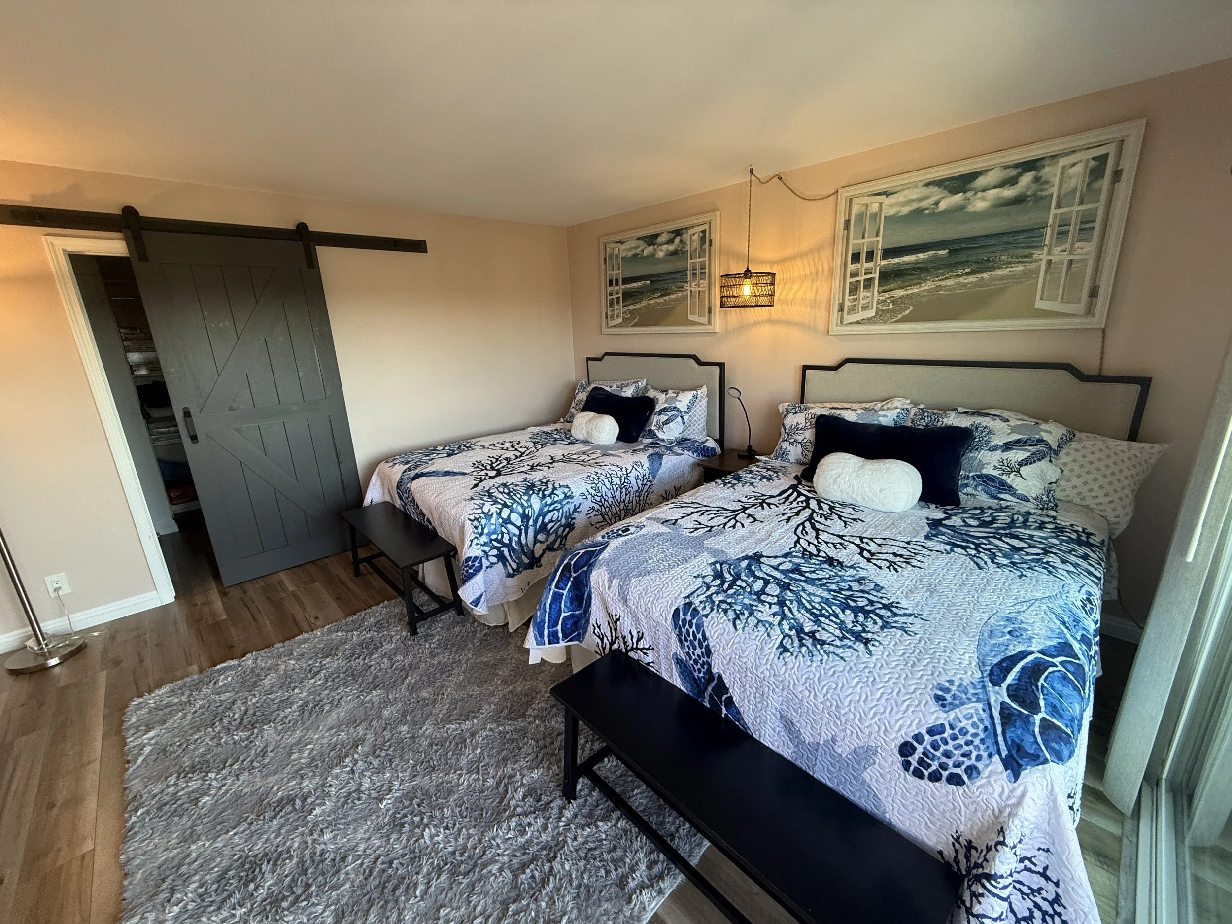 The Queens Suite bedroom in our Solana Beach vacation rental. A bedroom with two comfy queen-sized beds, an en suite, a walk-in closet with games and beach supplies, a plush rug, lamps, a La-Z-Boy, and a sliding glass door leading to a private balcon