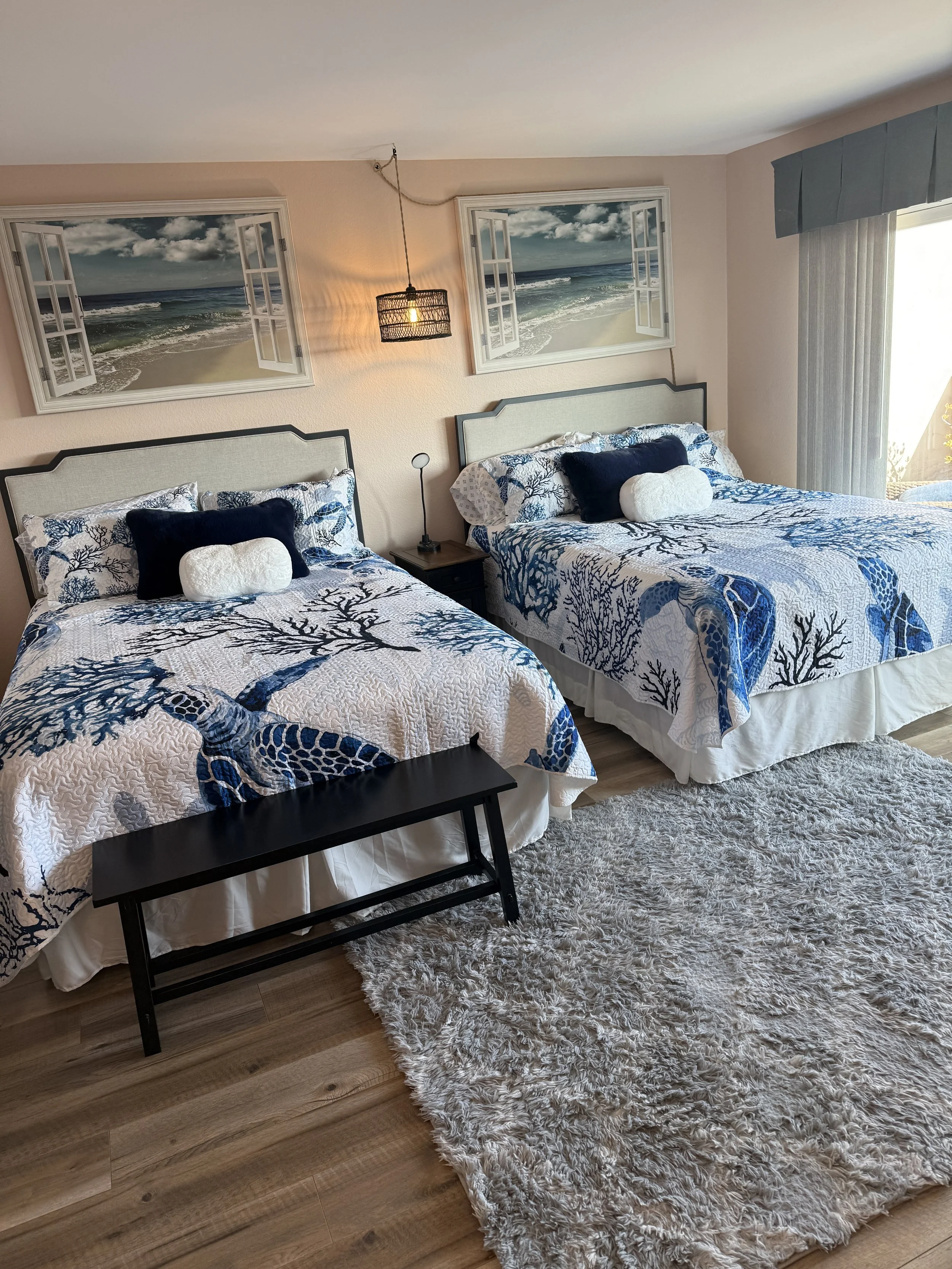 The Queens Suite bedroom in our Solana Beach vacation rental. A bedroom with two comfy queen-sized beds, an en suite, a walk-in closet with games and beach supplies, a plush rug, lamps, a La-Z-Boy, and a sliding glass door leading to a private balcon