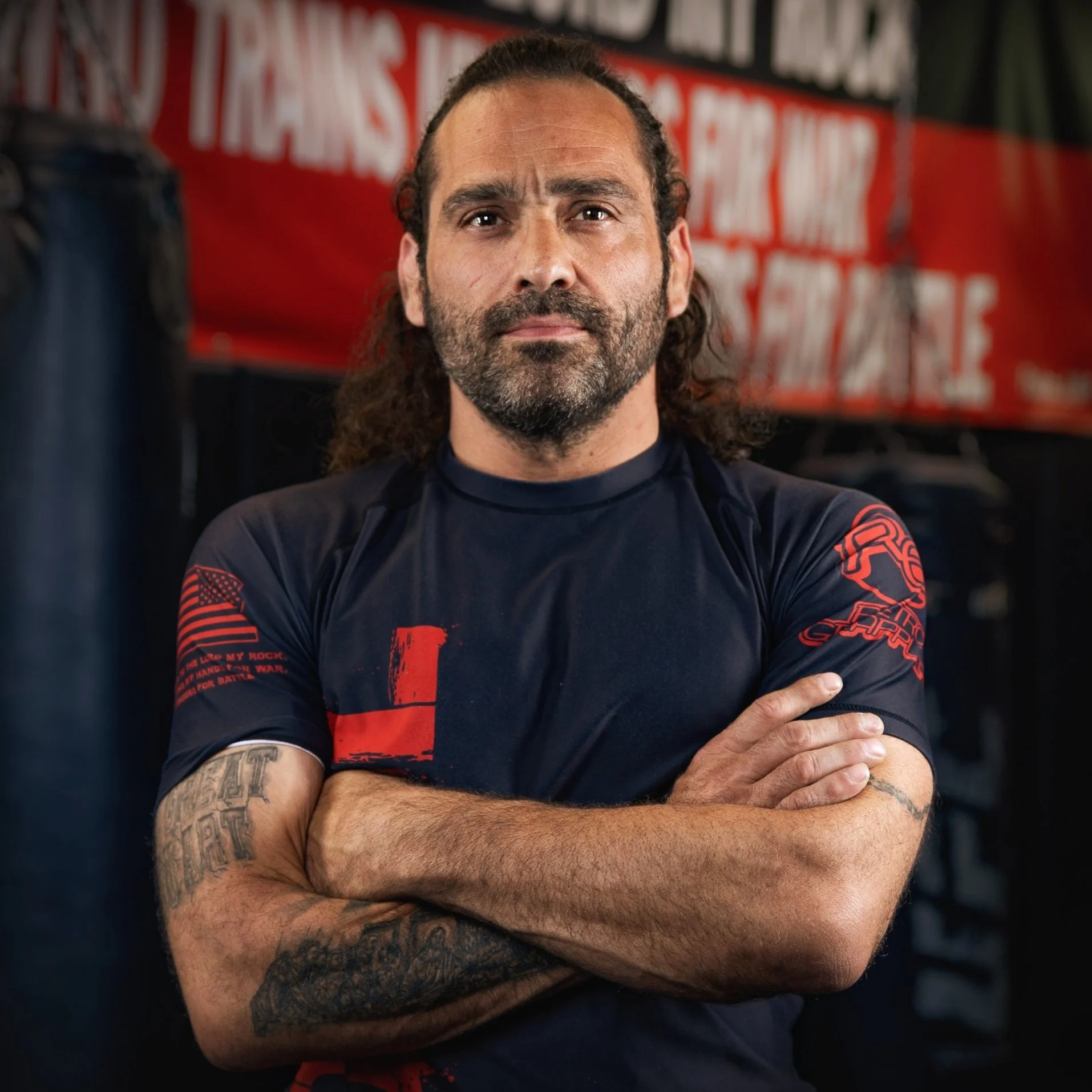 A man with long curly hair, a beard, and tattoos on his arms standing with arms crossed inside a gym, with a blurred red and black sign in the background.