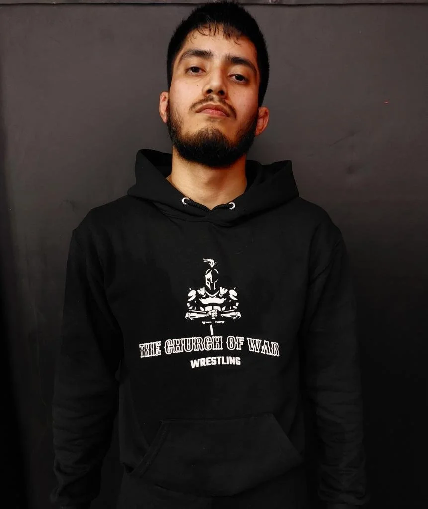 A young man with dark hair and a beard, wearing a black hoodie with the text 'The Church of War Wrestling' and a graphic of a knight, standing against a dark background.