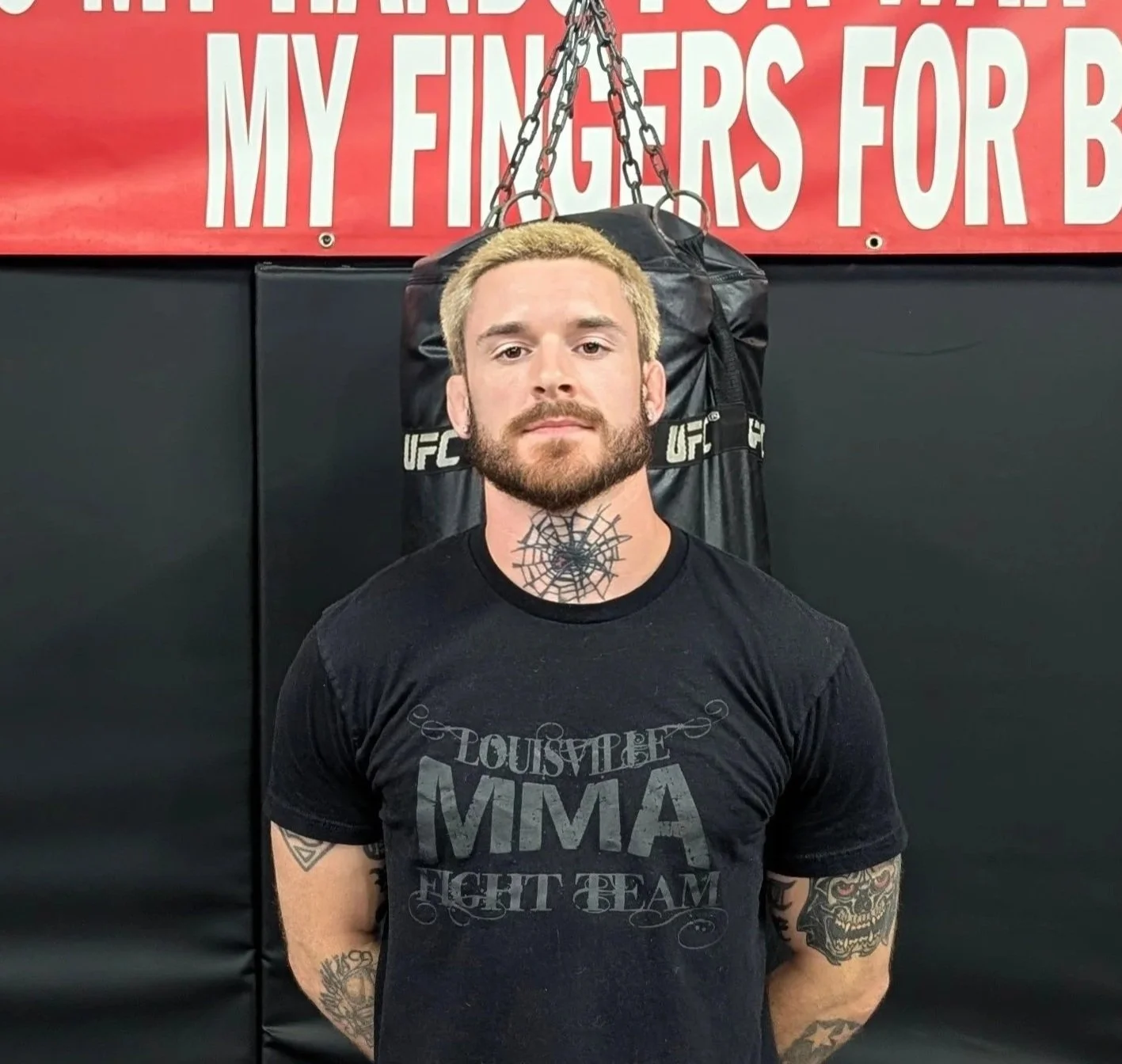 A shirtless male mixed martial arts fighter standing in front of a backdrop with MMA and Hardrock logos, wearing Under Armour shorts.