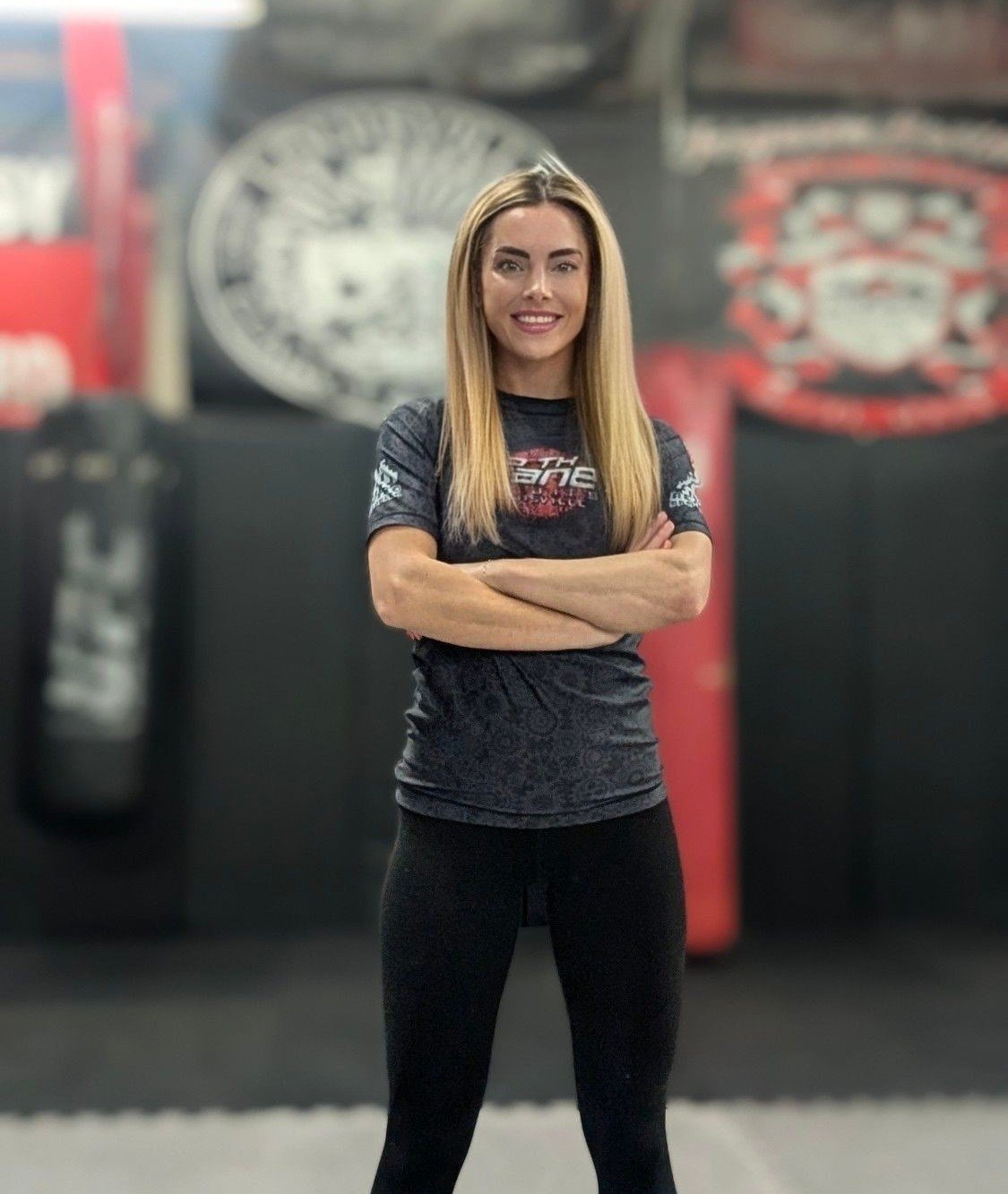 A woman standing with arms crossed in a gym or martial arts training area, smiling at the camera with boxing or kickboxing training equipment in the background.