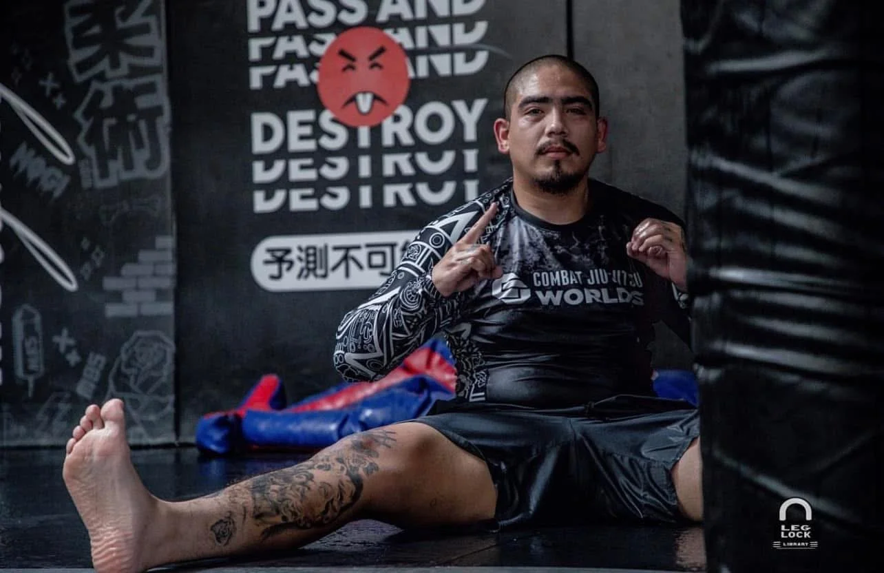 A man with a tattooed leg sitting on the floor in a martial arts gym, wearing a black and gray rash guard and shorts, giving a peace sign. Behind him, there is a black wall with martial arts posters, a red angry face emoji, and text that reads 'PASS AND DESTROY' and other phrases.