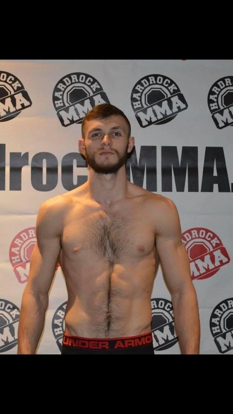 A shirtless male mixed martial arts fighter standing in front of a backdrop with MMA and Hardrock logos, wearing Under Armour shorts.