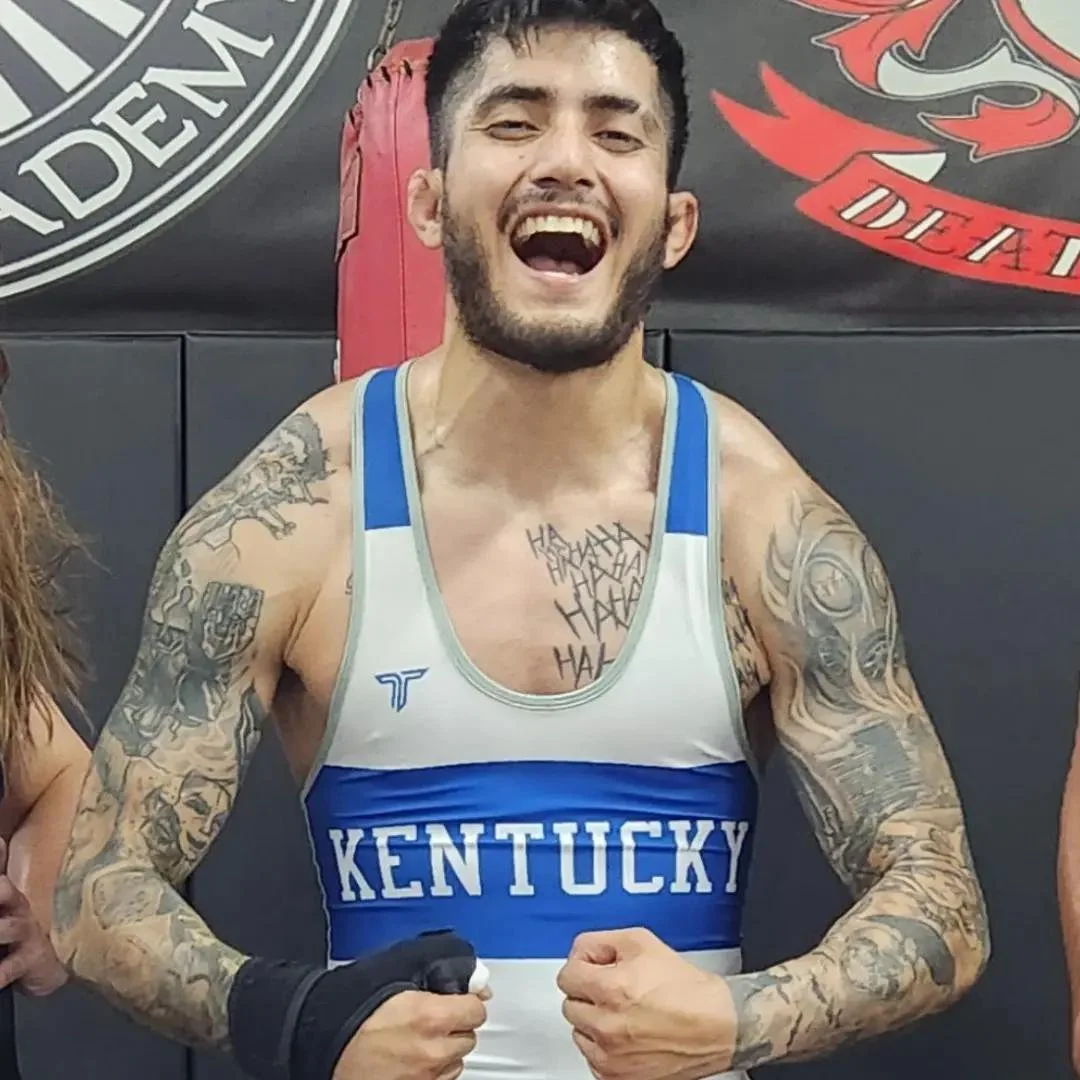 A young man with dark hair and a beard, wearing a black hoodie with the text 'The Church of War Wrestling' and a graphic of a knight, standing against a dark background.