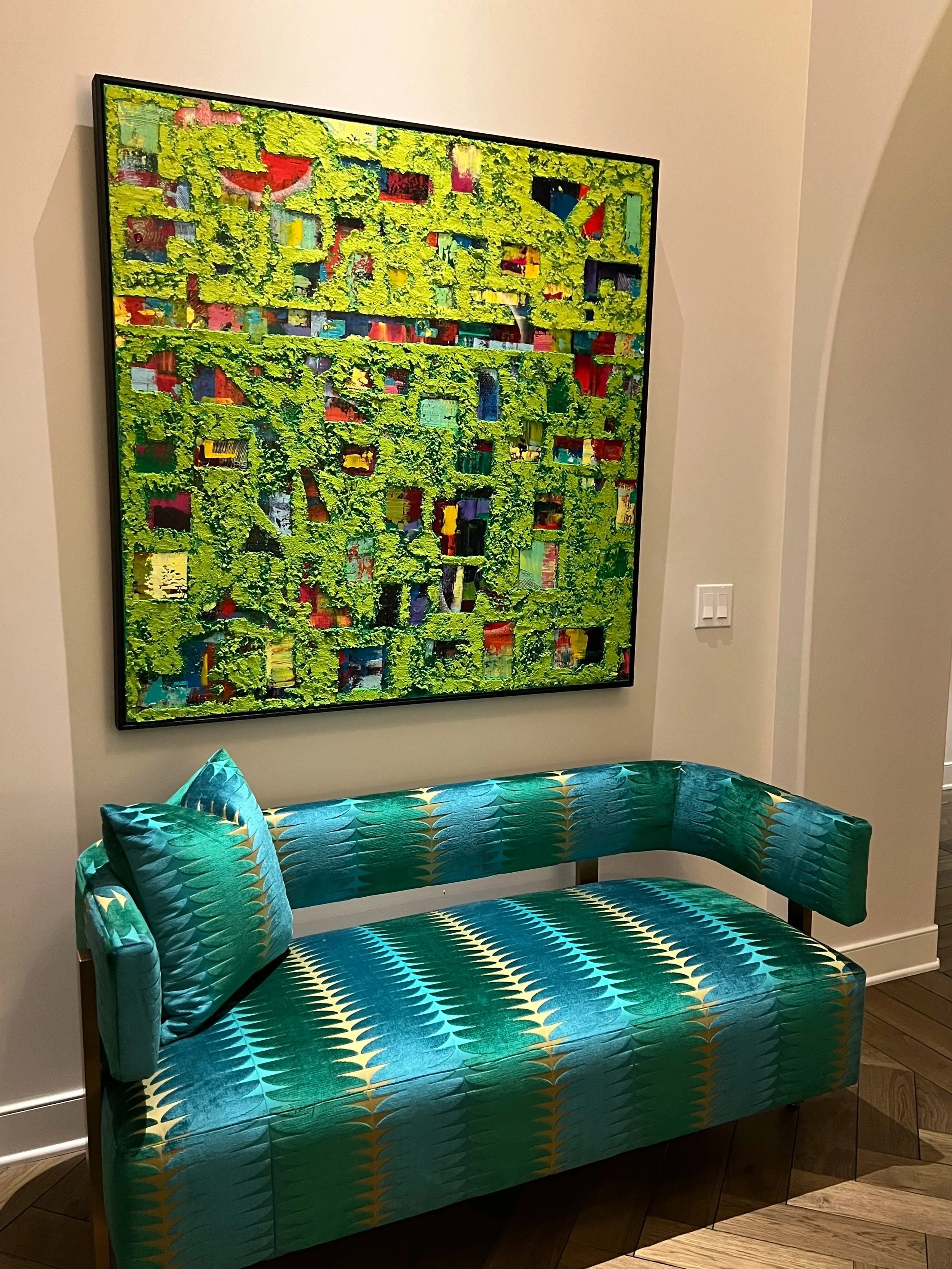 A vibrant teal and gold patterned settee with matching pillow in front of a colorful abstract painting on a beige wall.