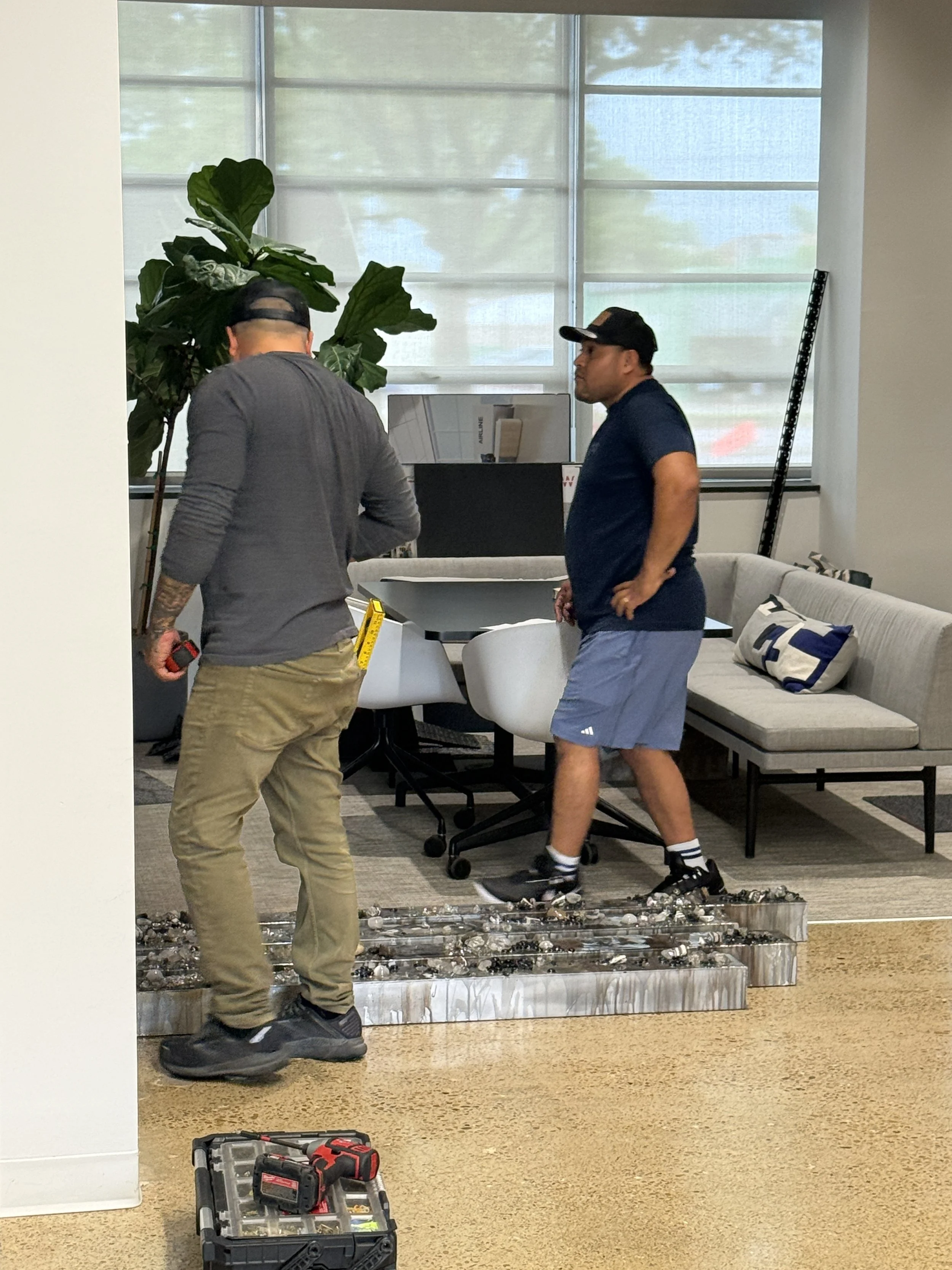 Two men working on a display or installation of small round objects, possibly sunglasses or similar items, in what appears to be an office or showroom. One man is holding a tool and headlamp, while the other stands nearby, wearing a baseball cap. There are tools and a toolbox on the floor, a large plant, and a window with blinds in the background.