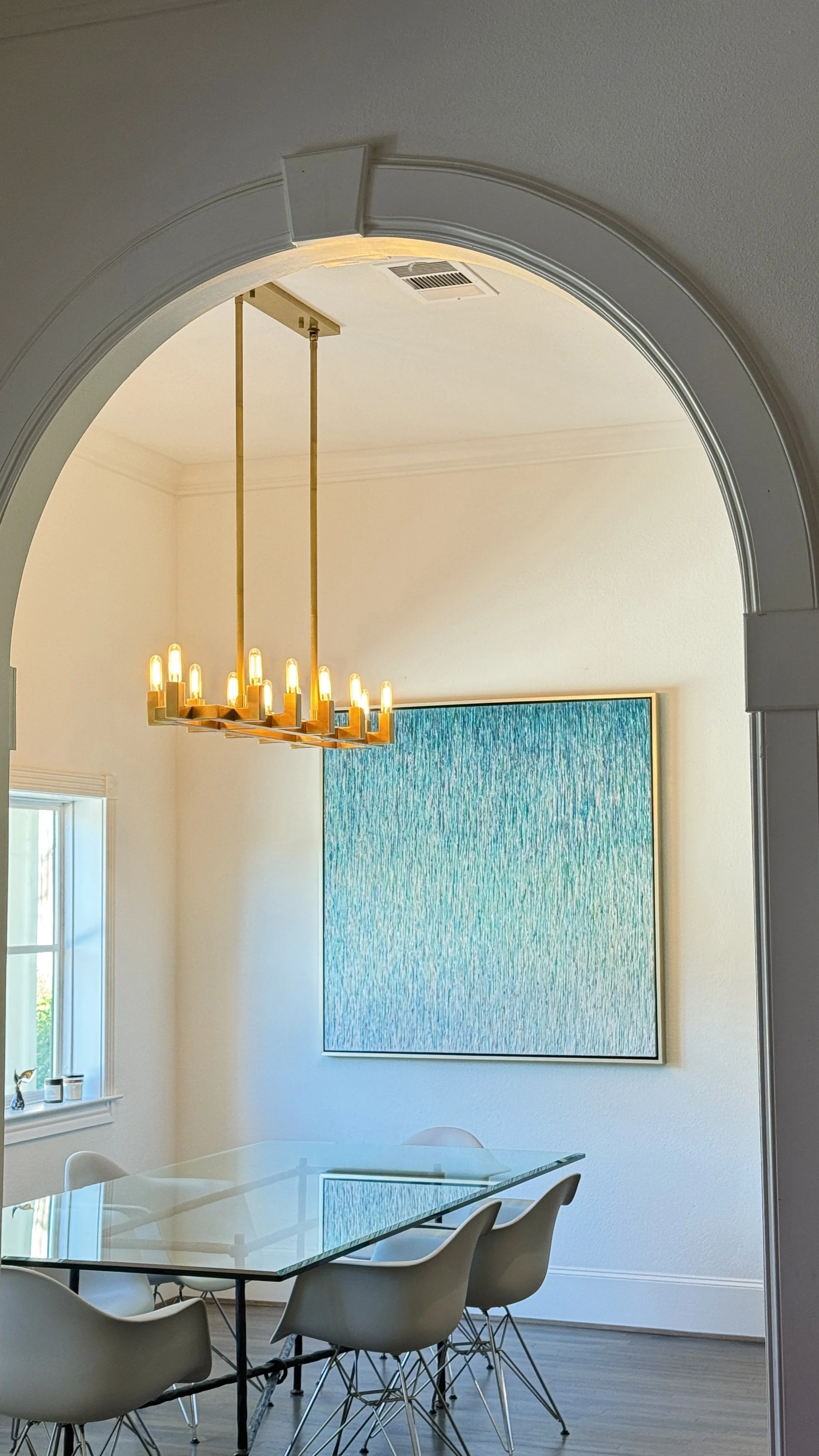 A dining room with a glass table, white chairs, a large blue textured artwork on the wall, a gold modern chandelier, a window with a small cat figurine and candles on the windowsill, and a doorway with an archway.
