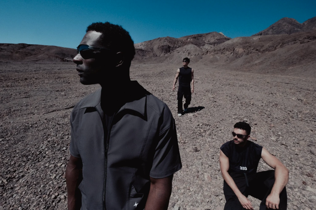 Three men in a rocky, desert landscape with mountains and a clear blue sky; one in the foreground wearing sunglasses and a gray vest, two others in the background, also wearing sunglasses, in dark clothing.
