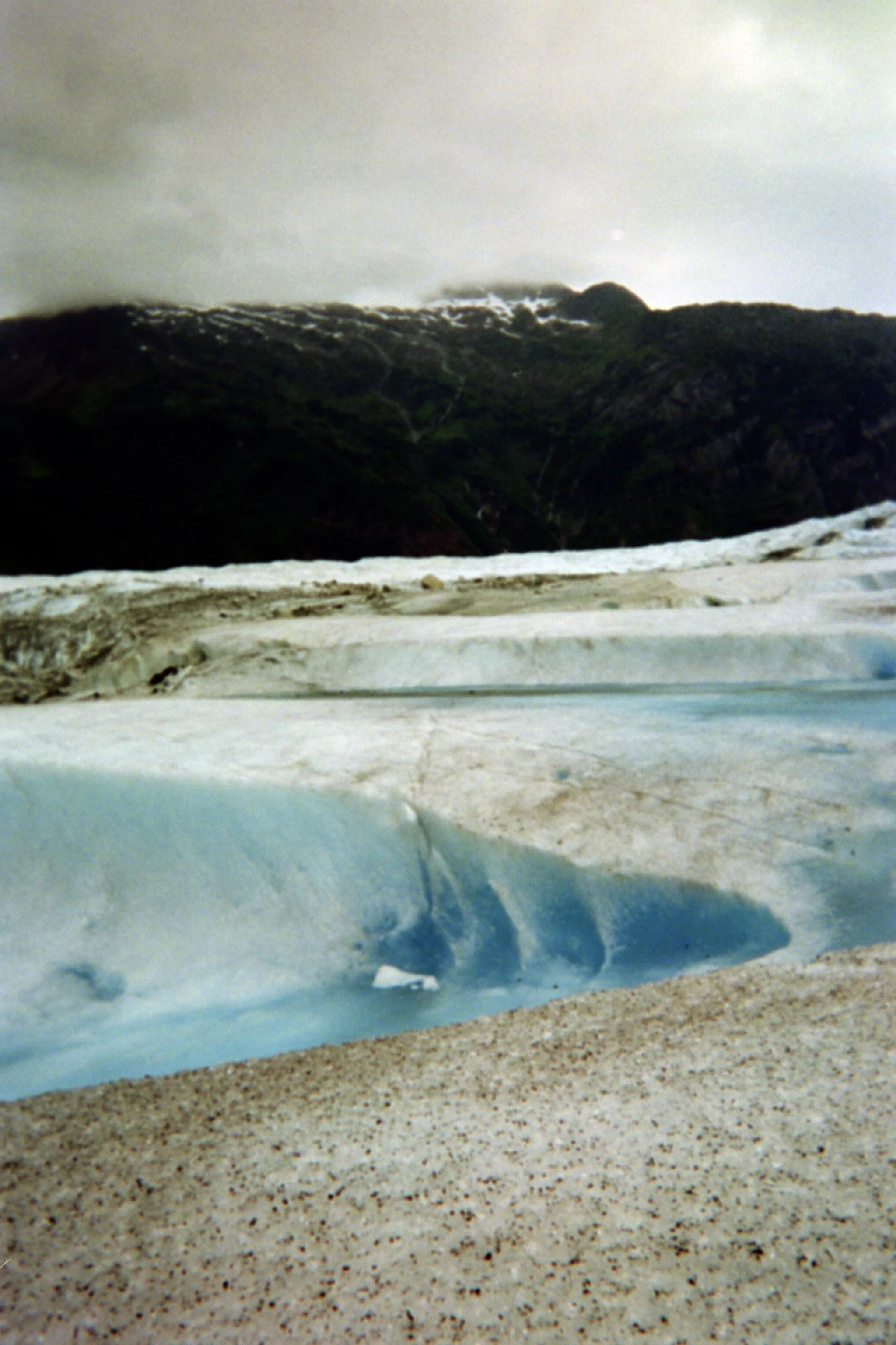 Close-up of a glacier with crack lines and ice blue color, with a mountain covered in snow and dark clouds in the background.
