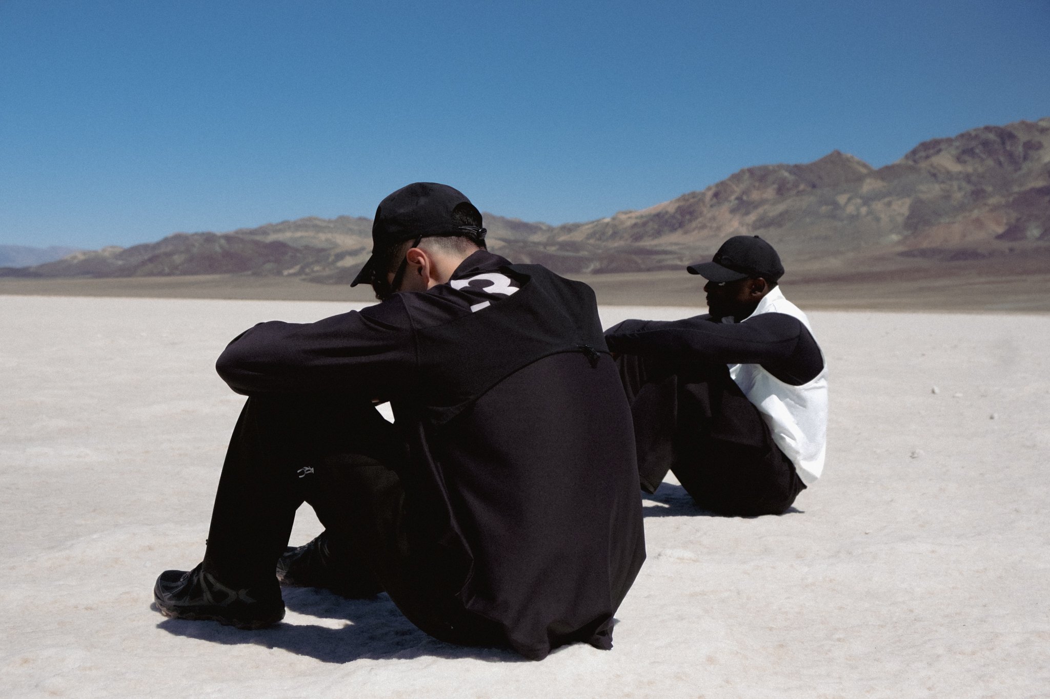 Two young men sitting on a bright white salt flat, with mountains and a clear blue sky in the background, one wearing a black cap and jacket, the other in a white and black jacket.