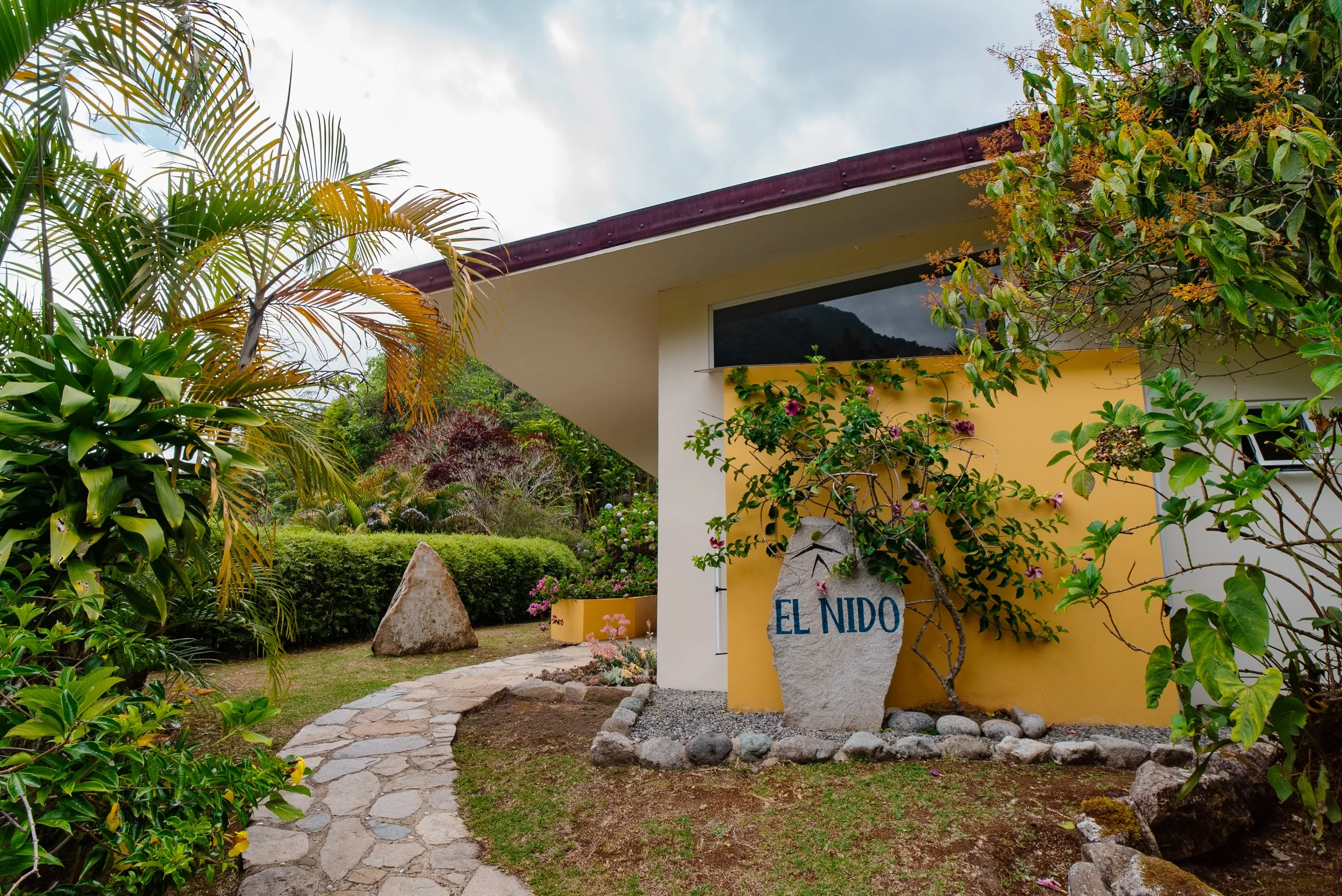 Casa pintada de amarillo con jardín y camino de piedra, rodeada de plantas y árboles, señal que dice 'El Nido'.