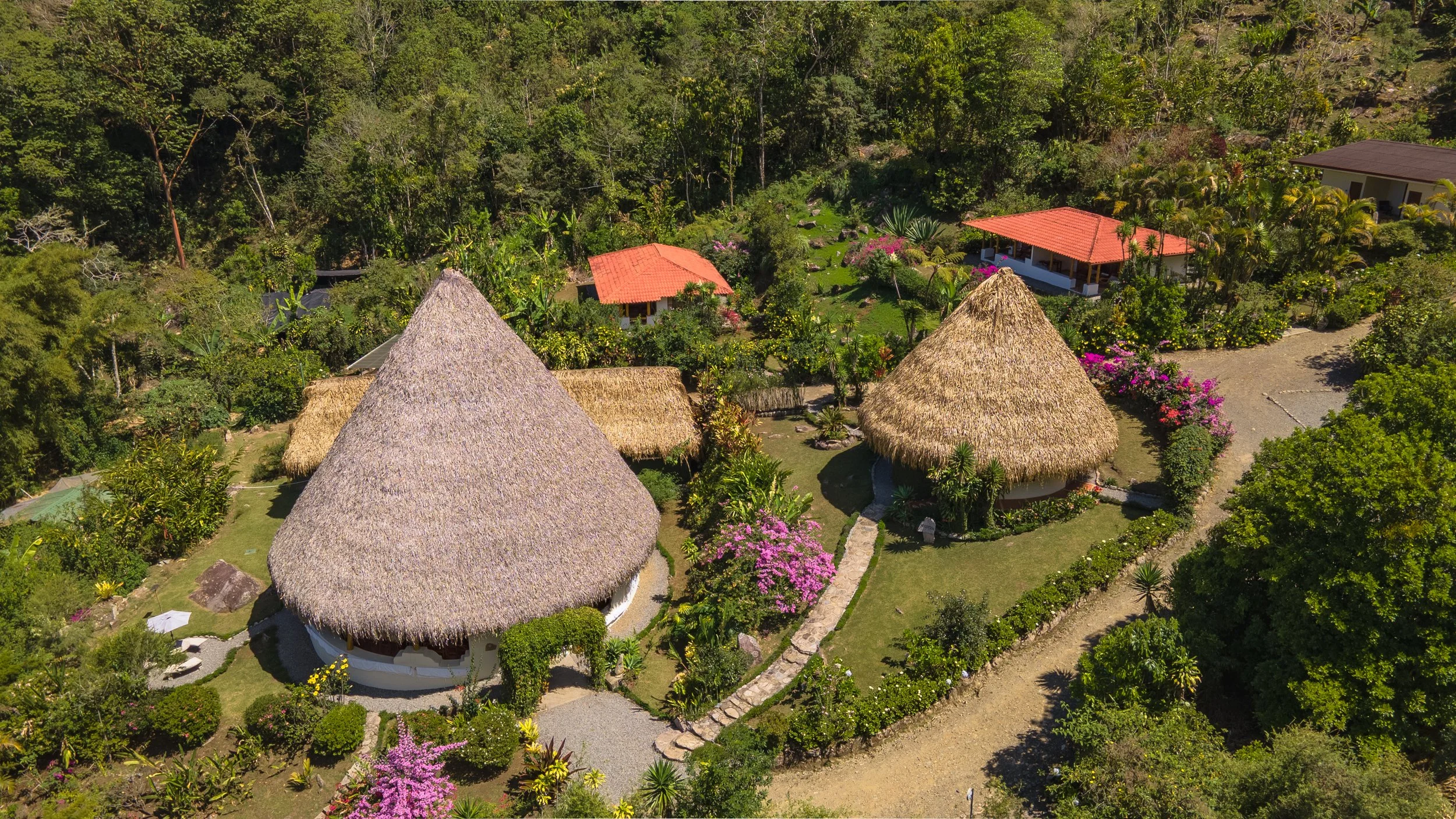 Vista aérea de un complejo de cabañas con techos de palma y jardines rodeados de vegetación y flores coloridas, en un entorno tropical.