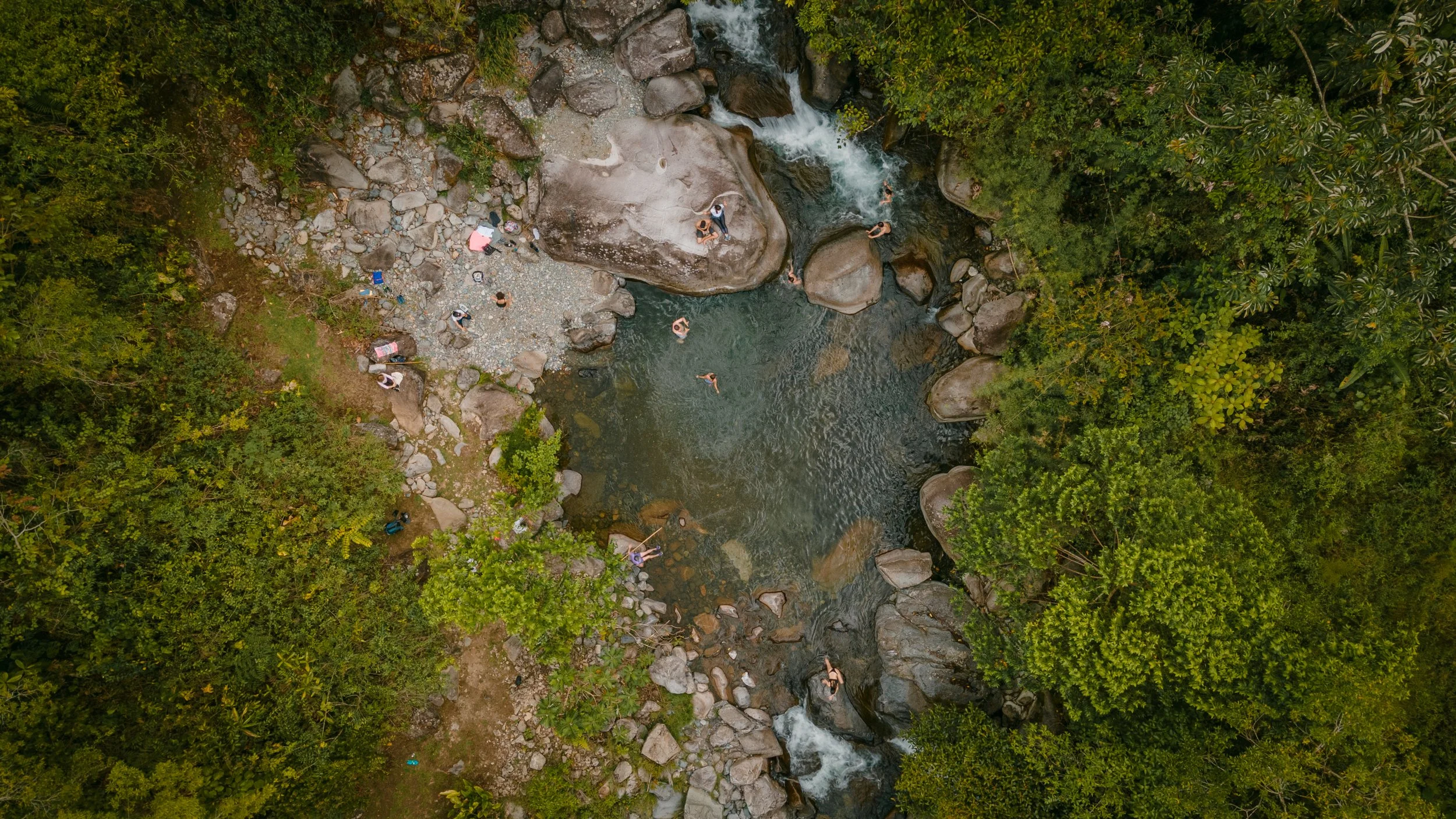 Persona en un río rodeado de naturaleza y rocas, algunos nadando y otros en la orilla, en un entorno natural con árboles verdes.