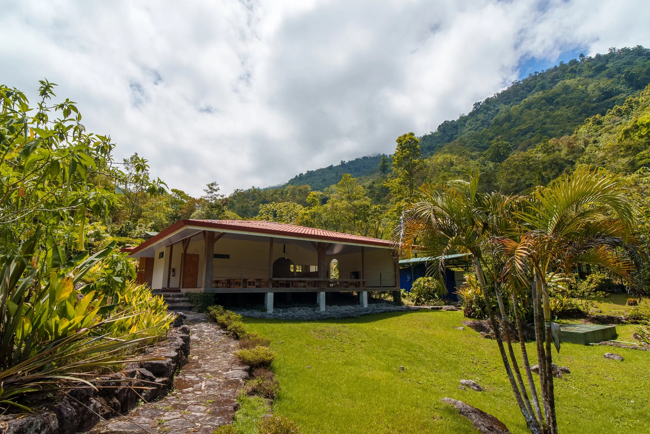 Casa en un entorno natural rodeada de árboles y montañas con cielo parcialmente nublado.
