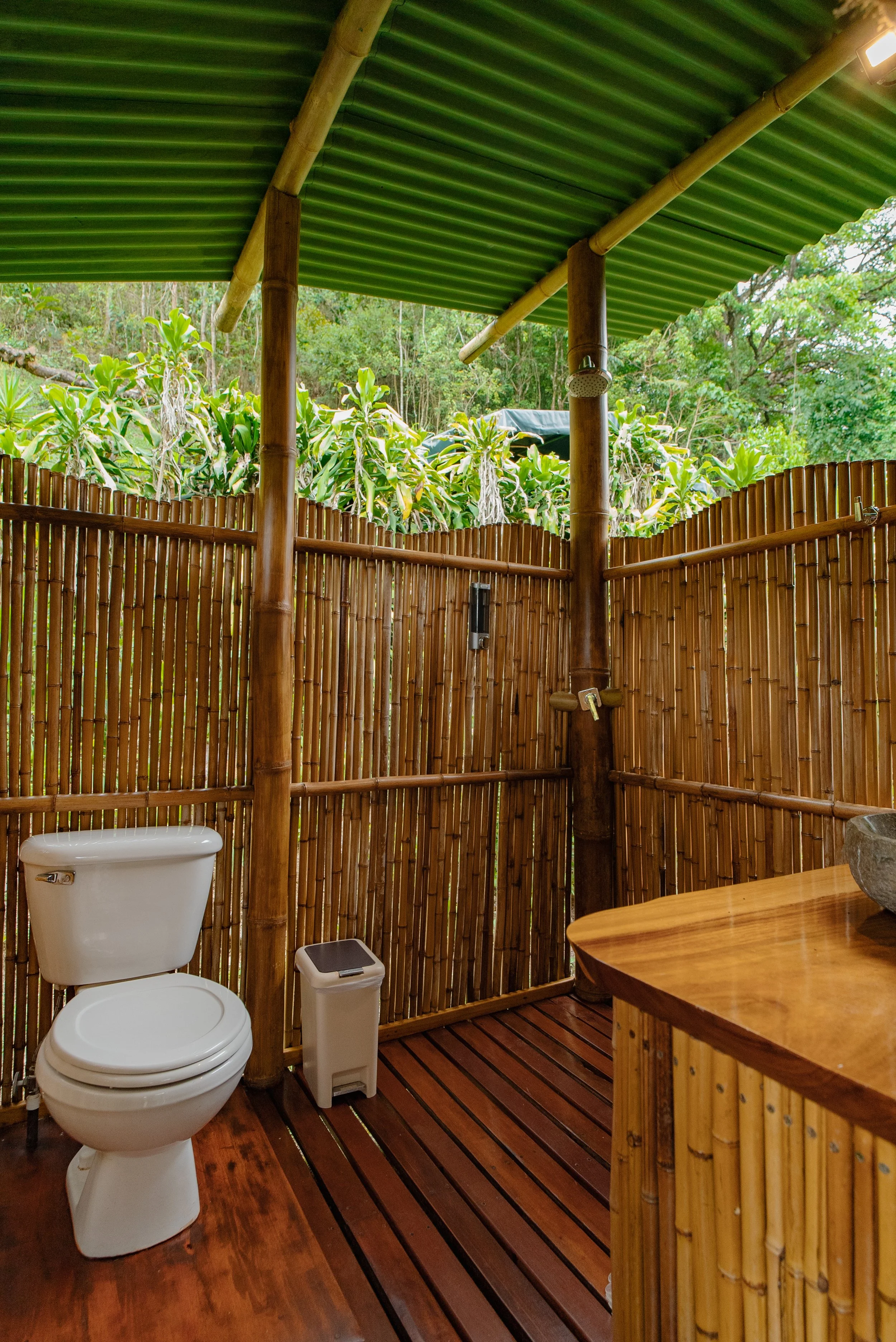 Baño ecológico con paredes de bambú, techo de láminas verdes y vista a la naturaleza, con inodoro, papelera y lavabo de piedra.