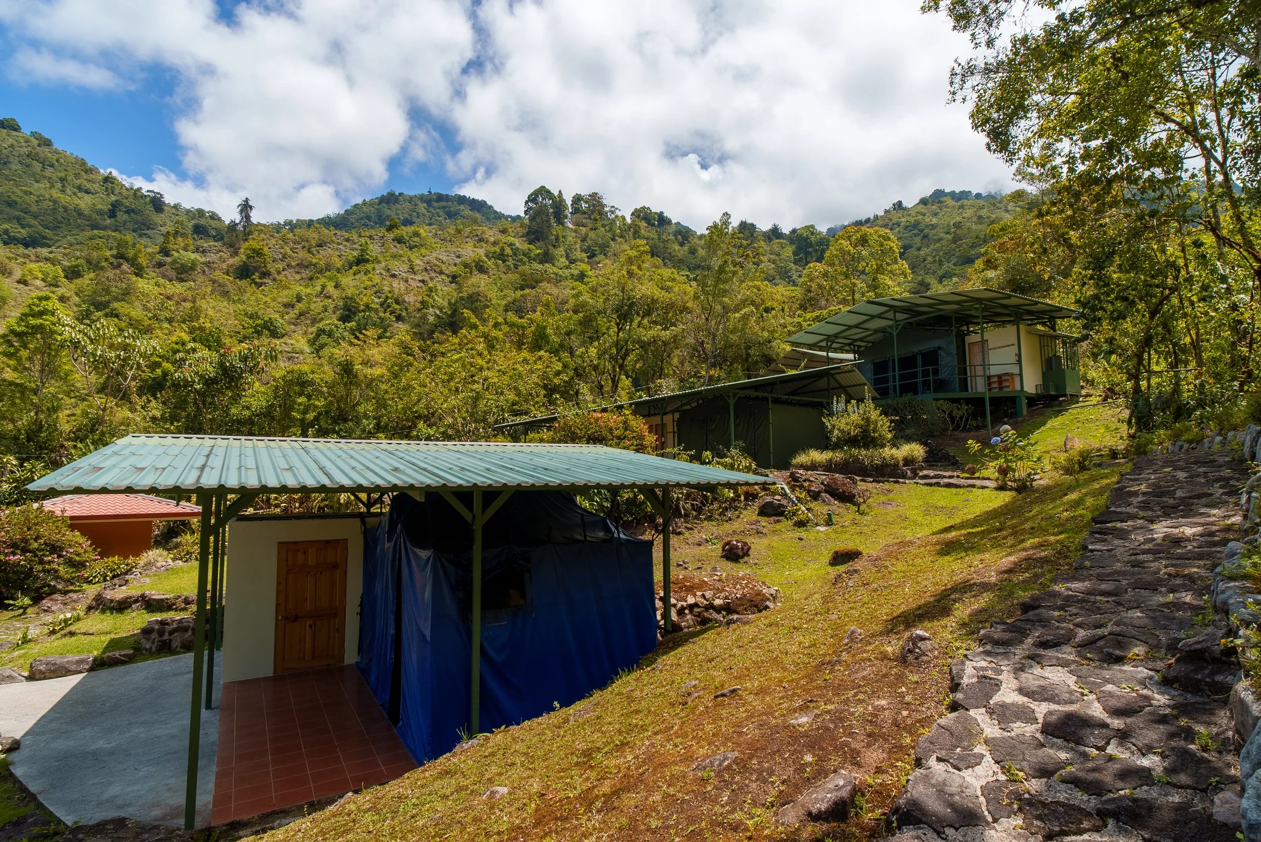 Casas de montaña en un paisaje natural con vegetación y colinas verdes, bajo un cielo parcialmente nublado.