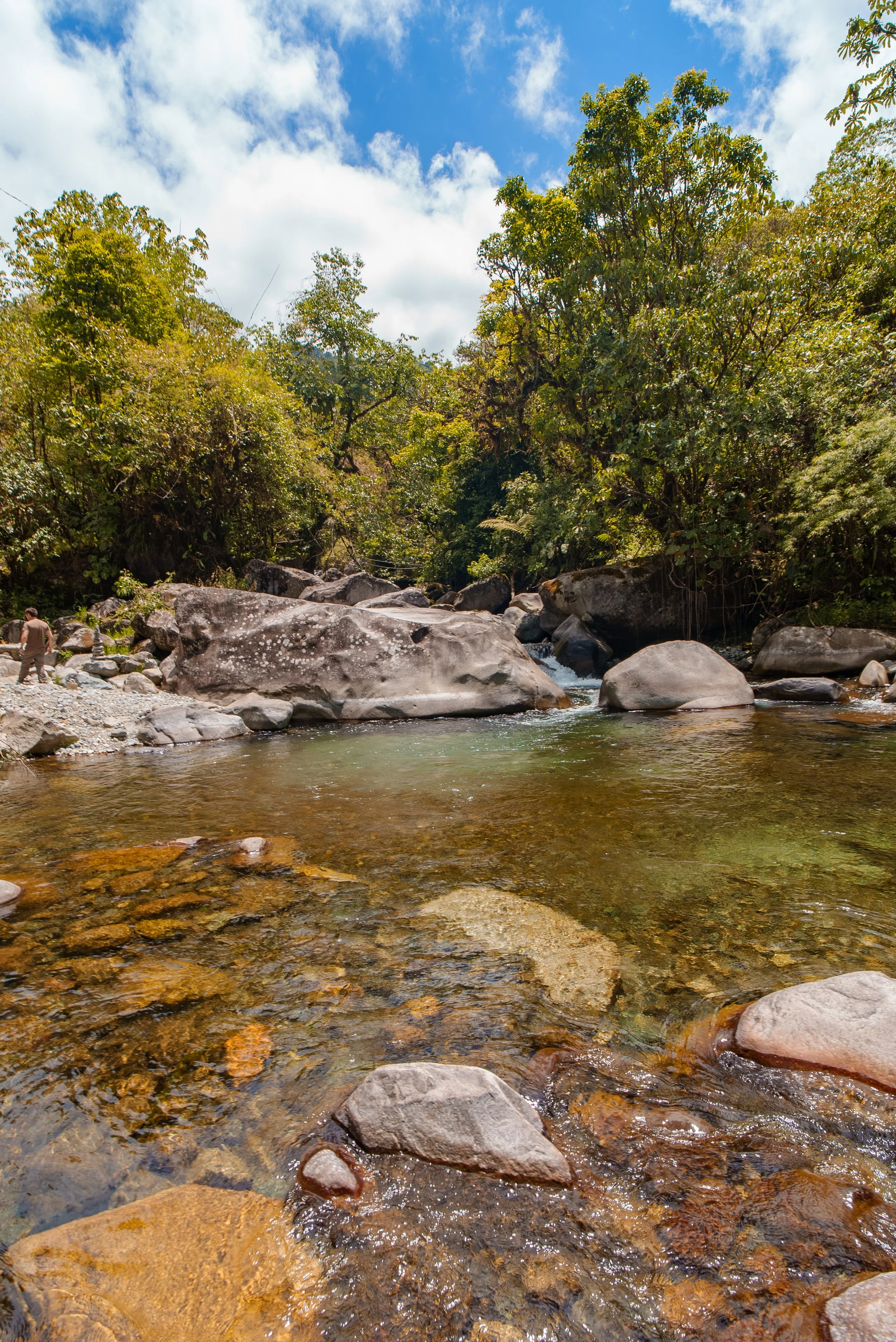 Un río cristalino rodeado de vegetación y rocas grandes en un día soleado con cielo azul y algunas nubes.